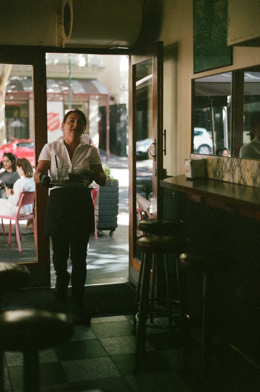 A woman in a white shirt holding a serving tray stands at the entrance of a restaurant, with outdoor seating and parked cars visible through the door.