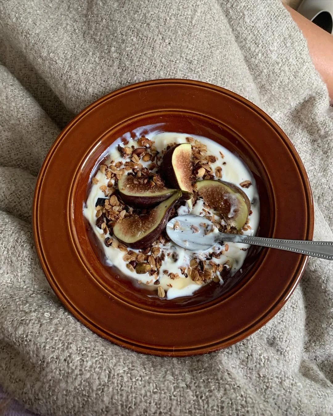 A brown bowl of yogurt topped with figs, granola, and honey, with a spoon in the bowl.