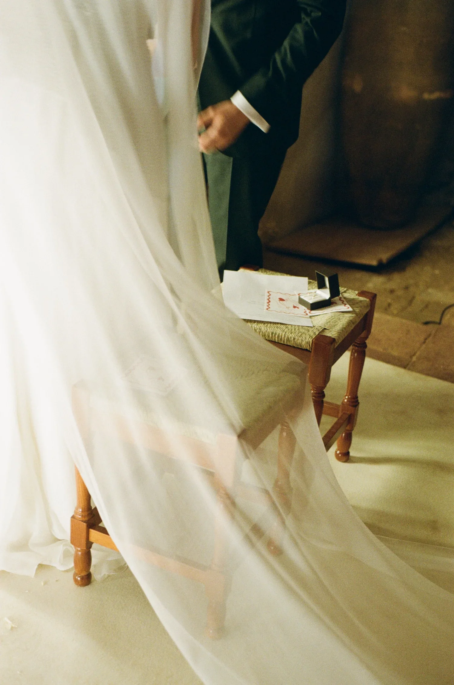 A bride in wedding gown with a veil, and a groom in a dark suit, standing next to a small wooden table with documents and a ring box.