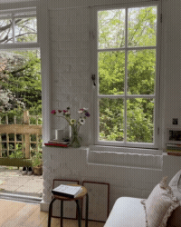 Bright room with a large window overlooking a green outdoor area, with a vase of flowers on a windowsill and a small stool with a book on it.