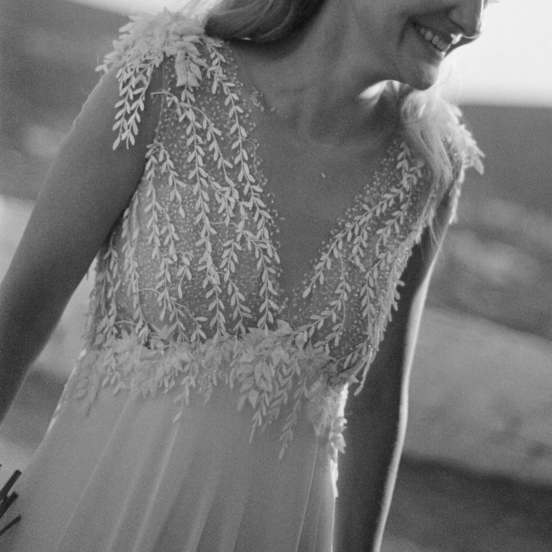 A woman in a wedding dress with lace and floral embroidery, smiling and leaning forward.