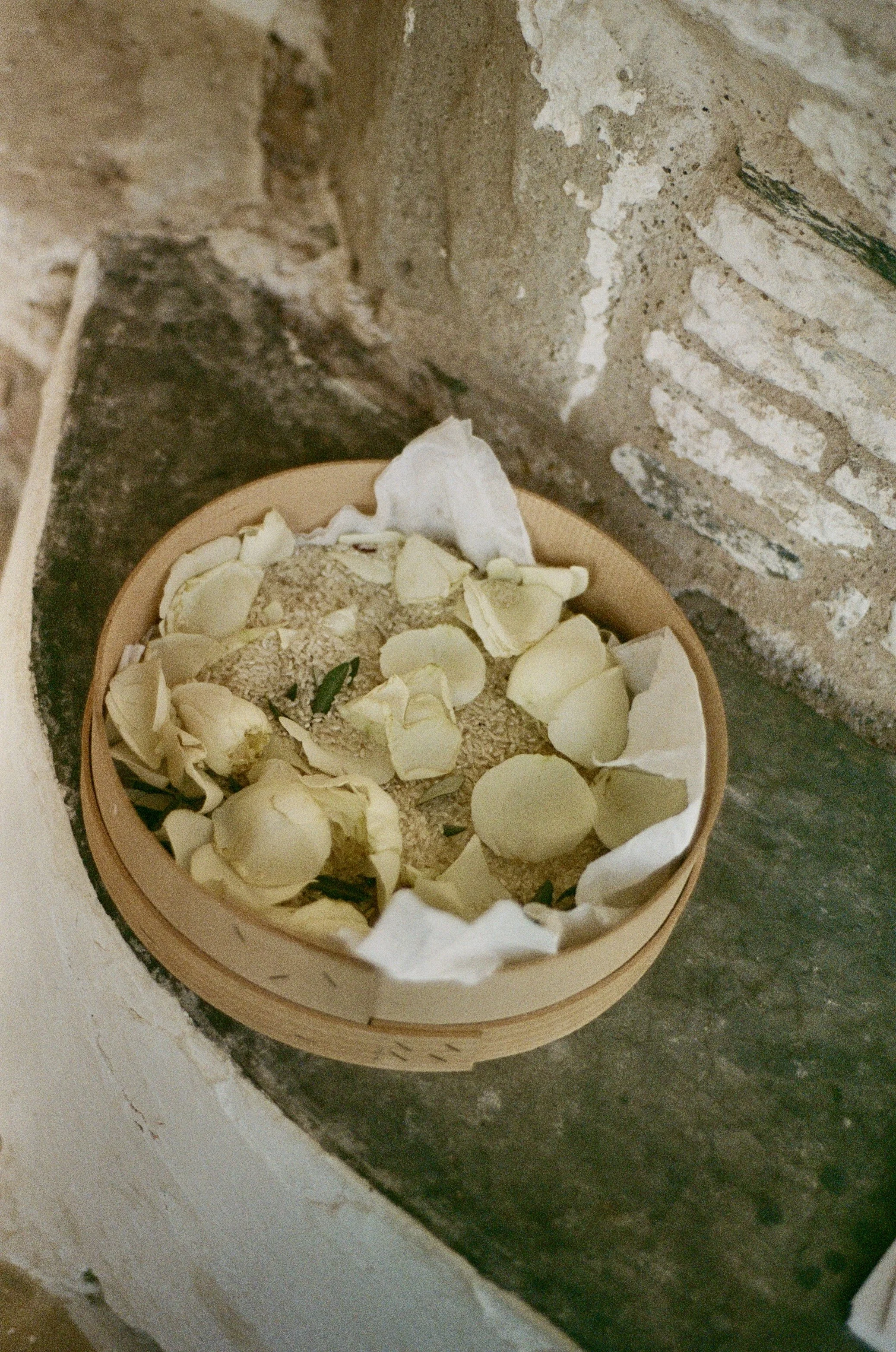 A small wooden dish with white flower petals on a dark stone surface next to a rustic brick wall.