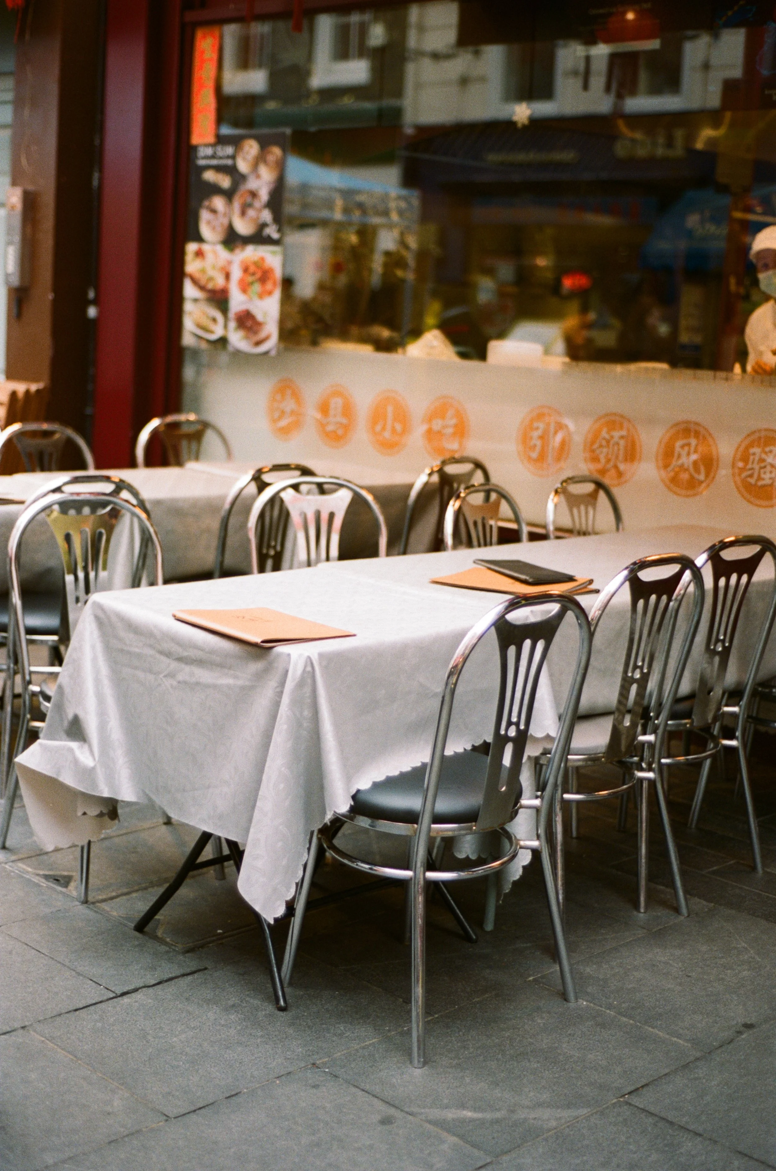 Empty outdoor restaurant seating with tables covered in white tablecloths and black and silver chairs, outside a Asian restaurant.