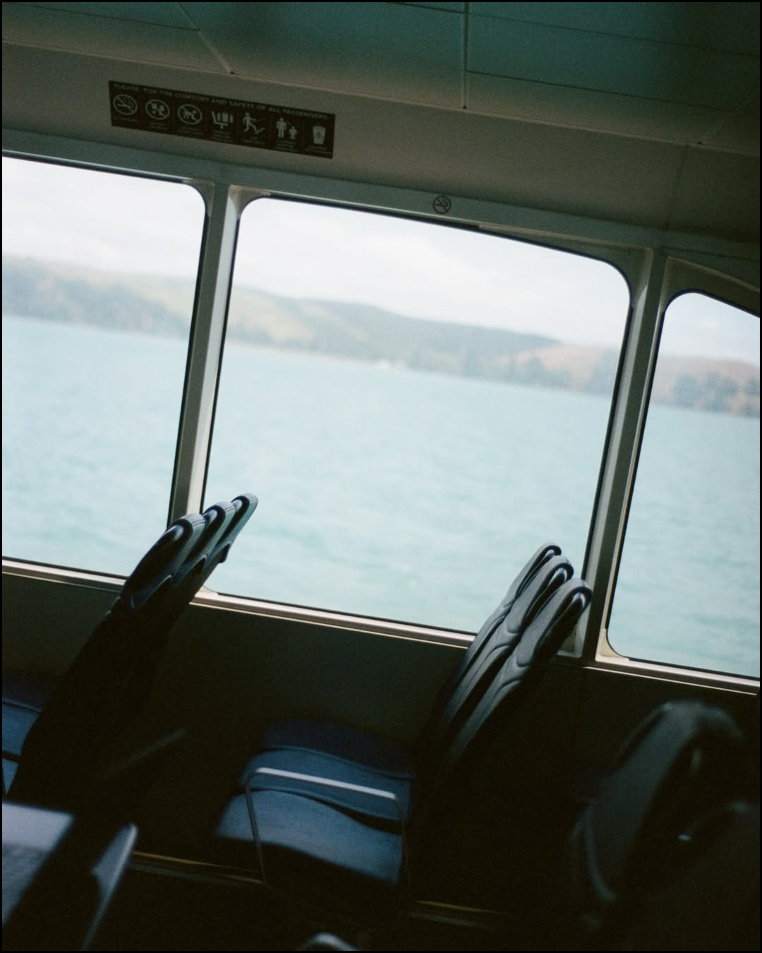 Inside a ferry boat with empty black seats and a large window showing a body of water and distant hills.