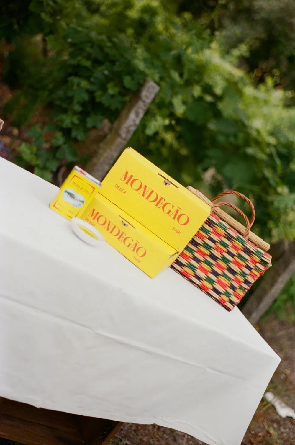 A table displaying yellow boxes labeled 'MONDEGÃO DESDE 1966', a small can, a small container, and colorful woven baskets, set outdoors with greenery in the background.
