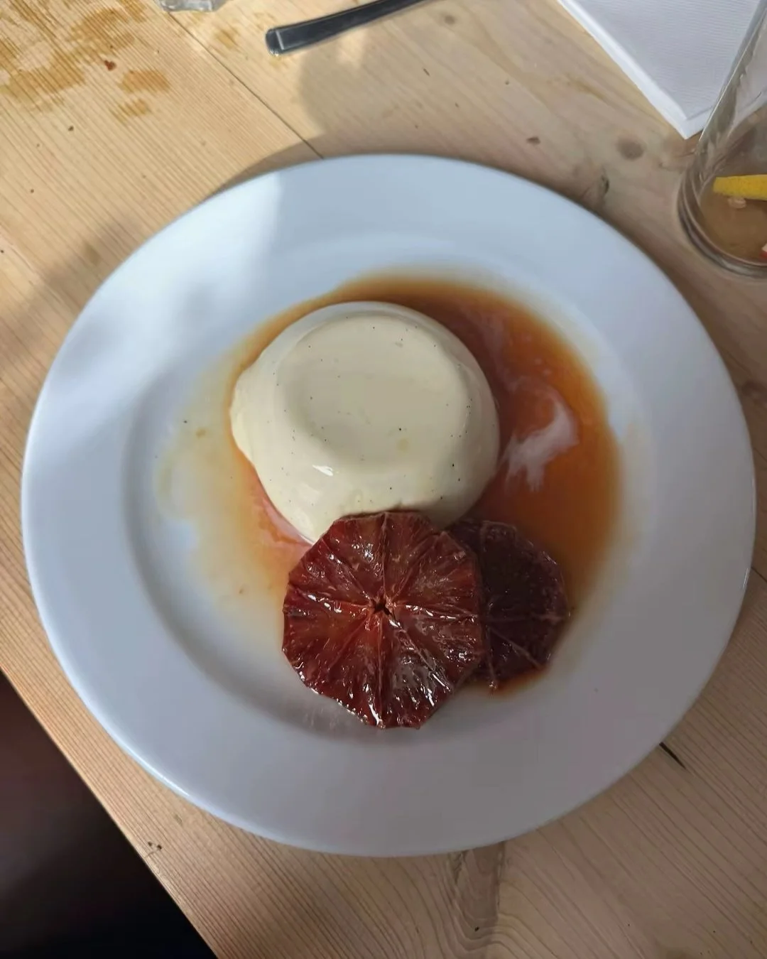 A white plate with a scoop of vanilla ice cream, a slice of blood orange, and caramel sauce drizzled around the ice cream, on a wooden table with a glass of lemon water and a pen nearby.