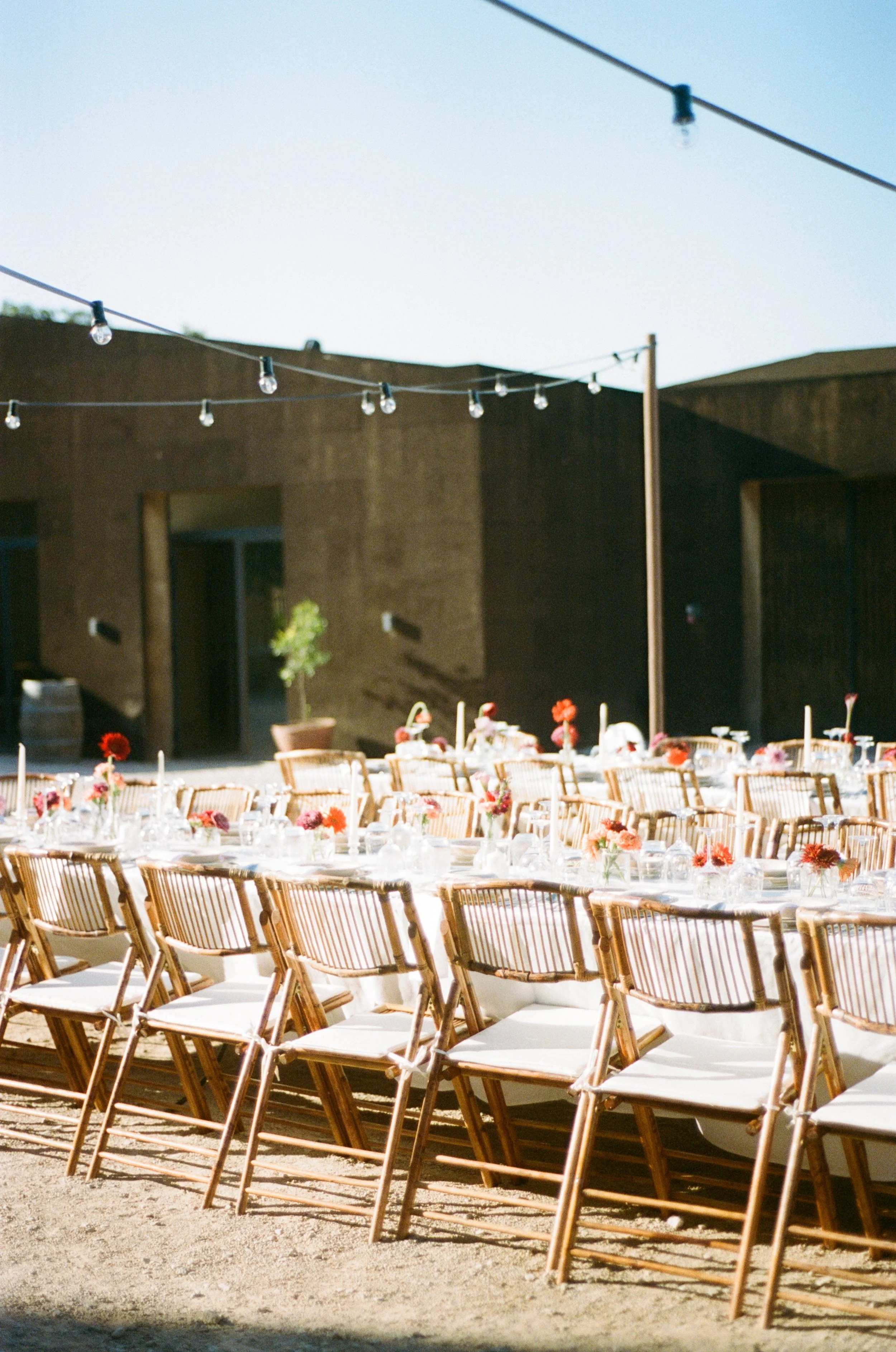 Outdoor dining setup with tables and chairs, decorated with small floral arrangements and candles, under string lights on a sunny day.