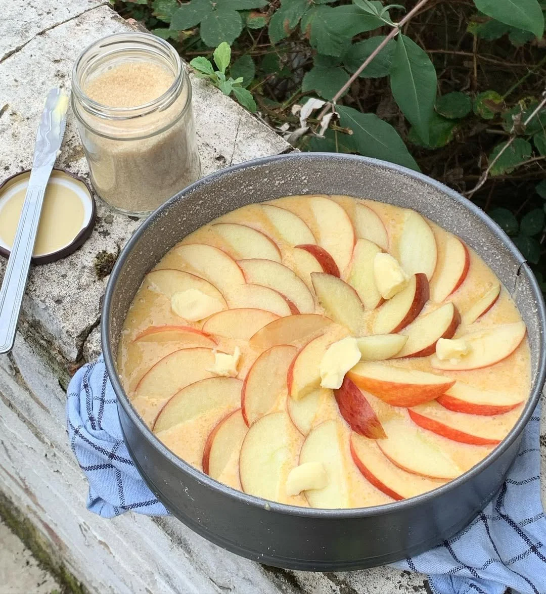 A baking dish filled with sliced apples and pieces of butter, ready to be baked, placed on a cloth on a stone surface outdoors, with a jar of sugar and a small dish of cream or sauce nearby.