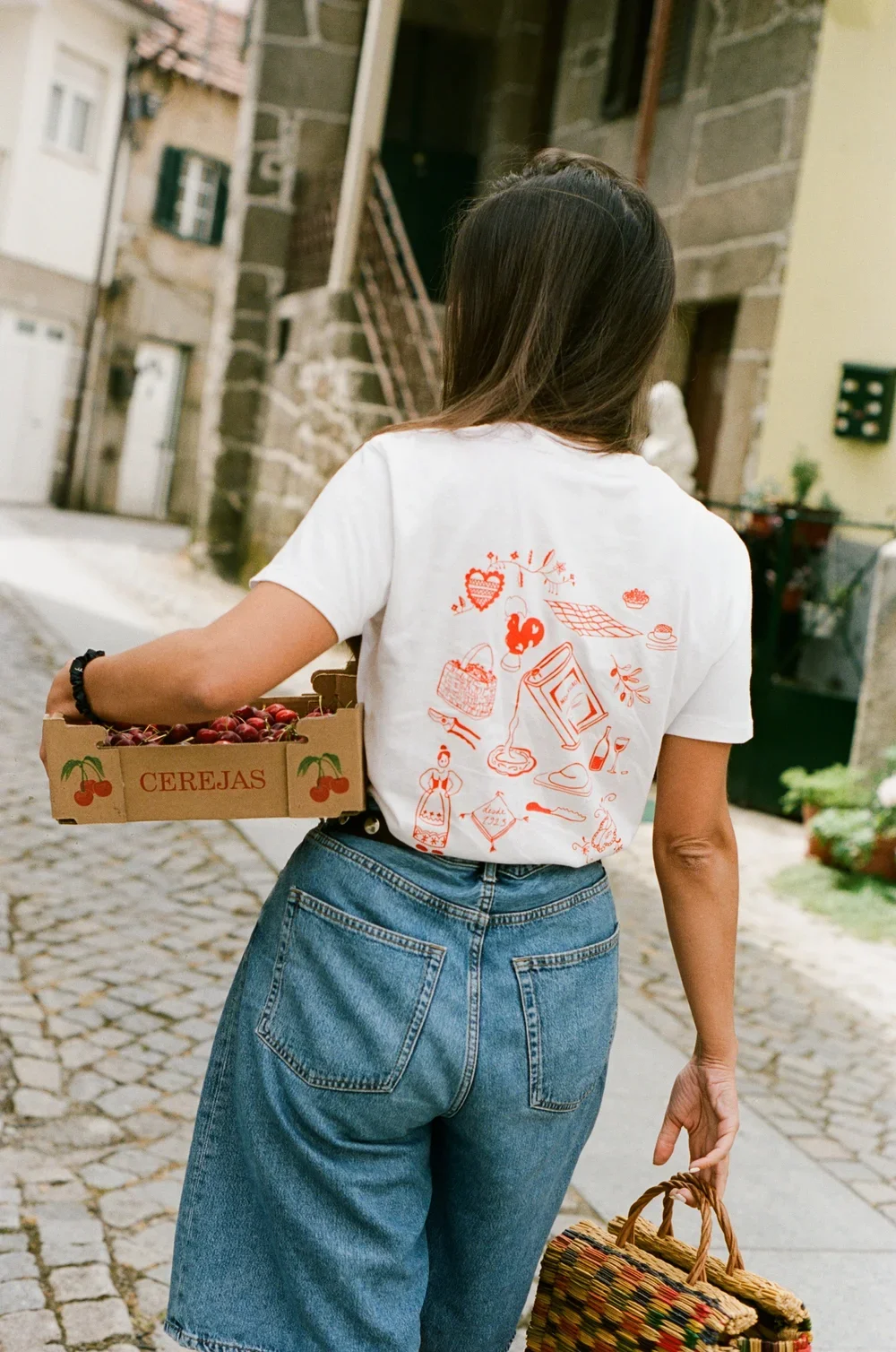 A woman walking down a cobblestone street carrying a cardboard box of cherries labeled 'CEREJAS' in her left hand, and a woven basket in her right hand, wearing a white T-shirt with orange illustrations on the back and blue jeans.