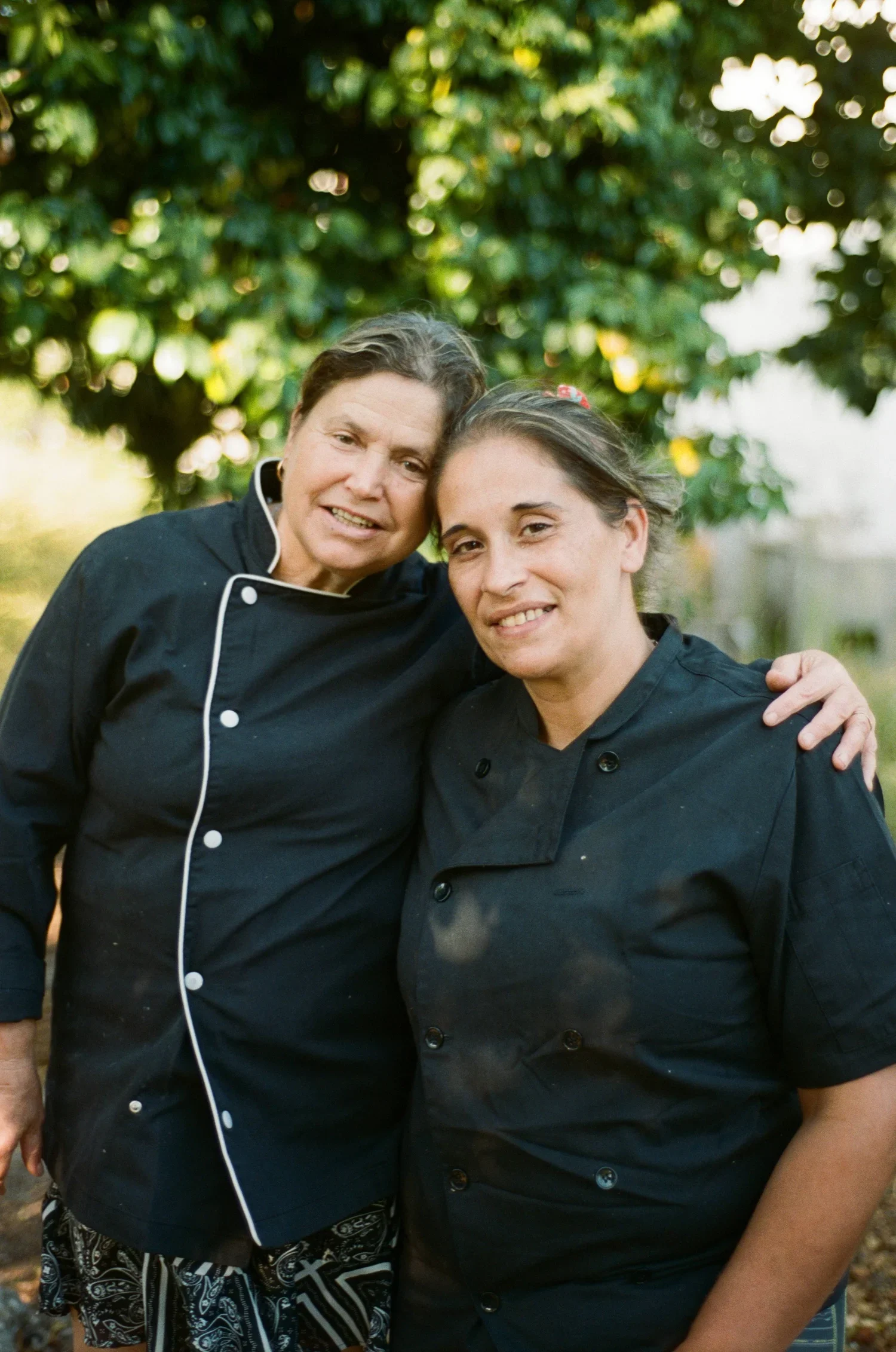 Two women in black chef coats posing outdoors in front of a leafy tree, smiling with their heads touching and arms around each other.