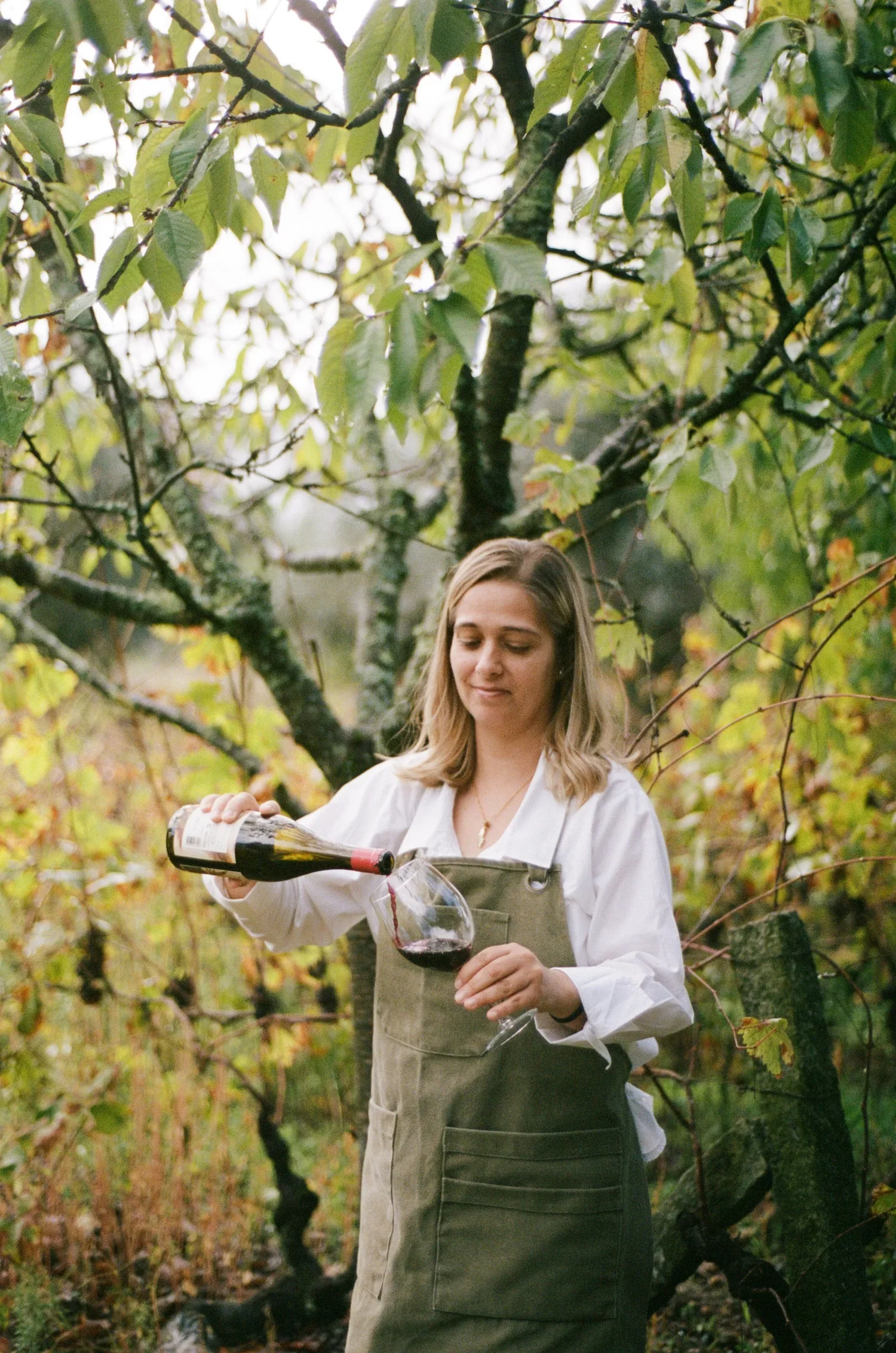 A woman in an outdoor setting holds a wine glass and pours red wine from a bottle with trees and greenery in the background.