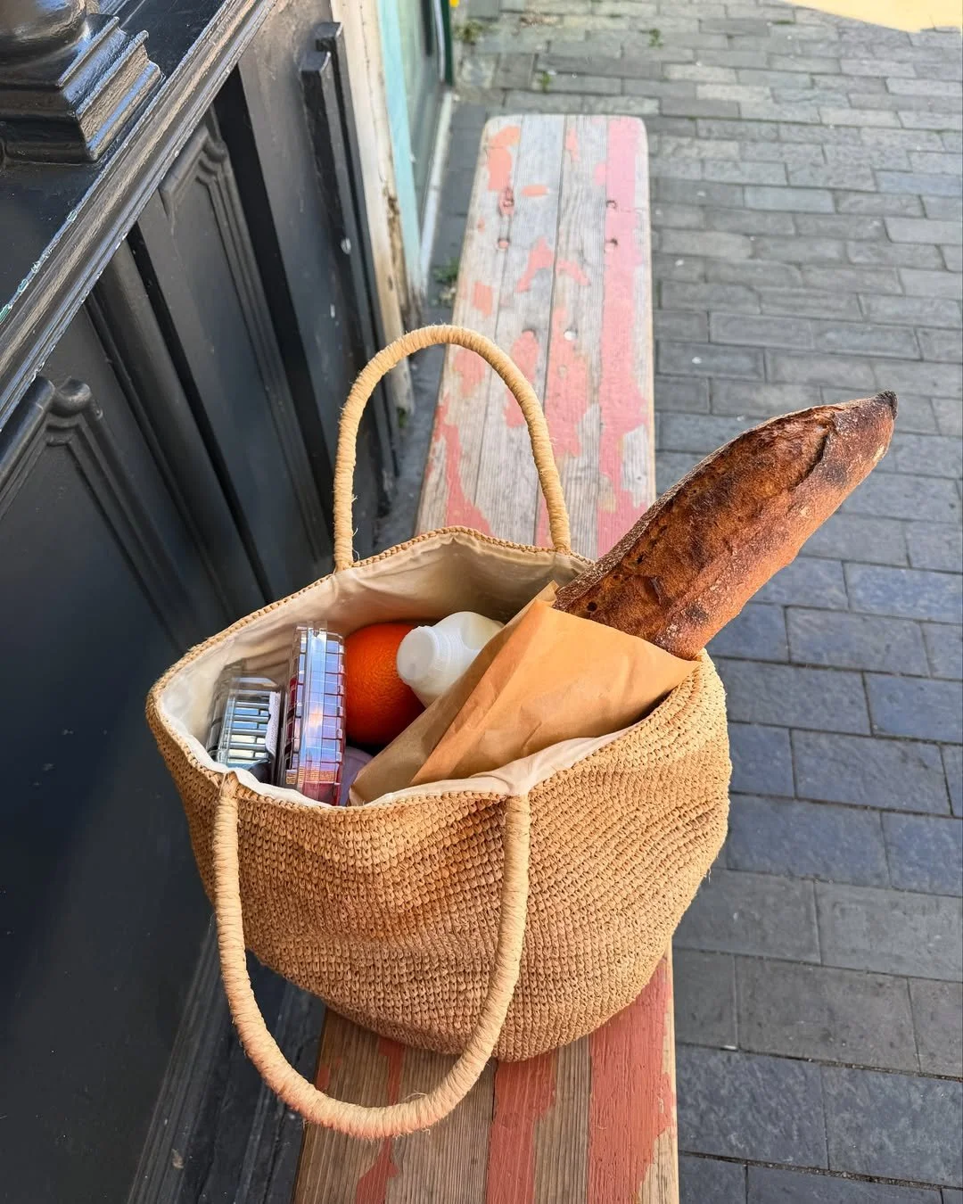 A woven beige tote bag with groceries, including a baguette, an orange, a plastic container, a small bottle, and a paper bag, resting on a worn wooden bench on a sidewalk.