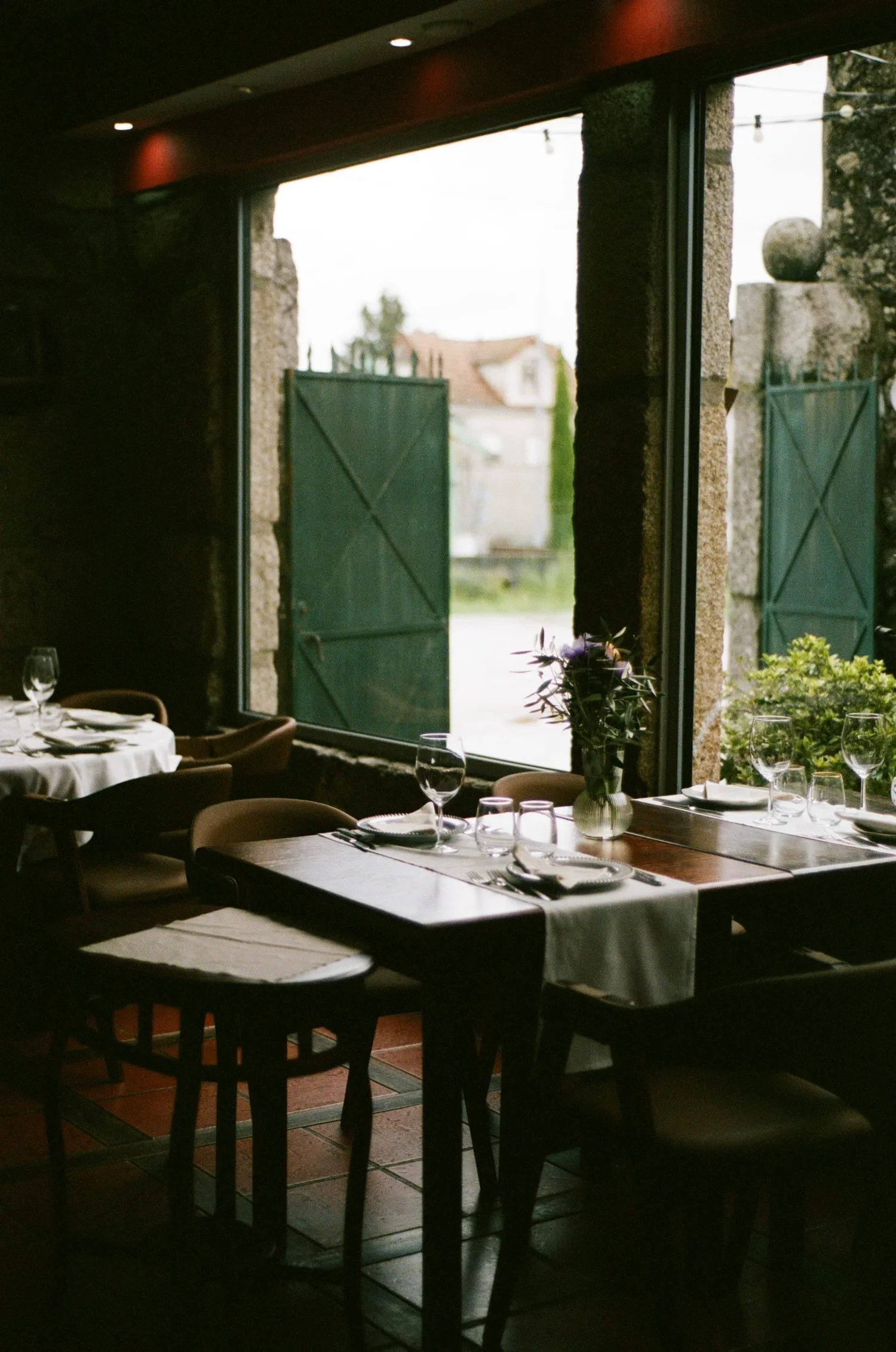 Interior of a restaurant with tables set with glasses, plates, and silverware, facing a large window with green shutters and outdoor scenery.