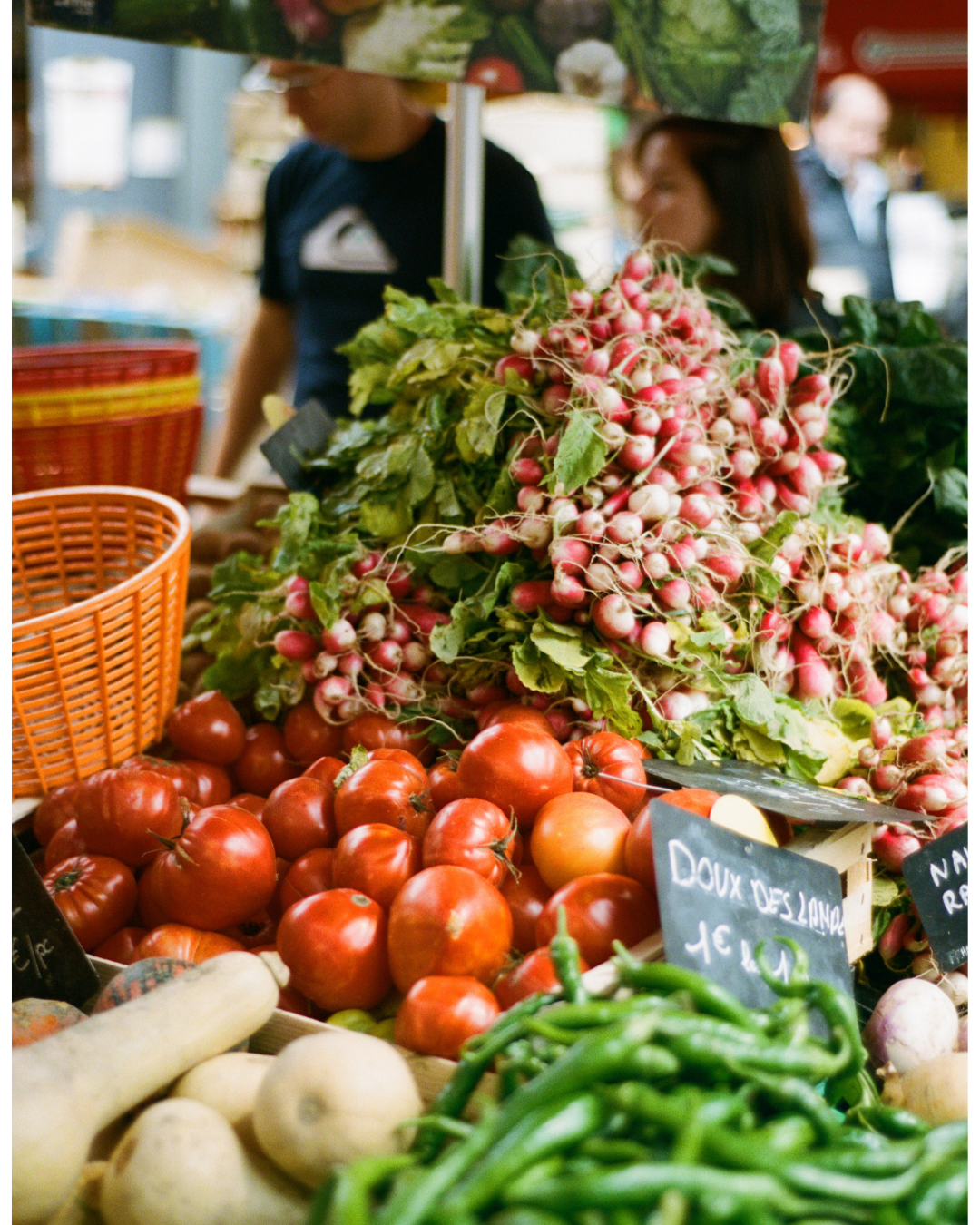 A display of fresh vegetables at a market, including tomatoes, radishes, green chili peppers, and white vegetables, with blurred people in the background.