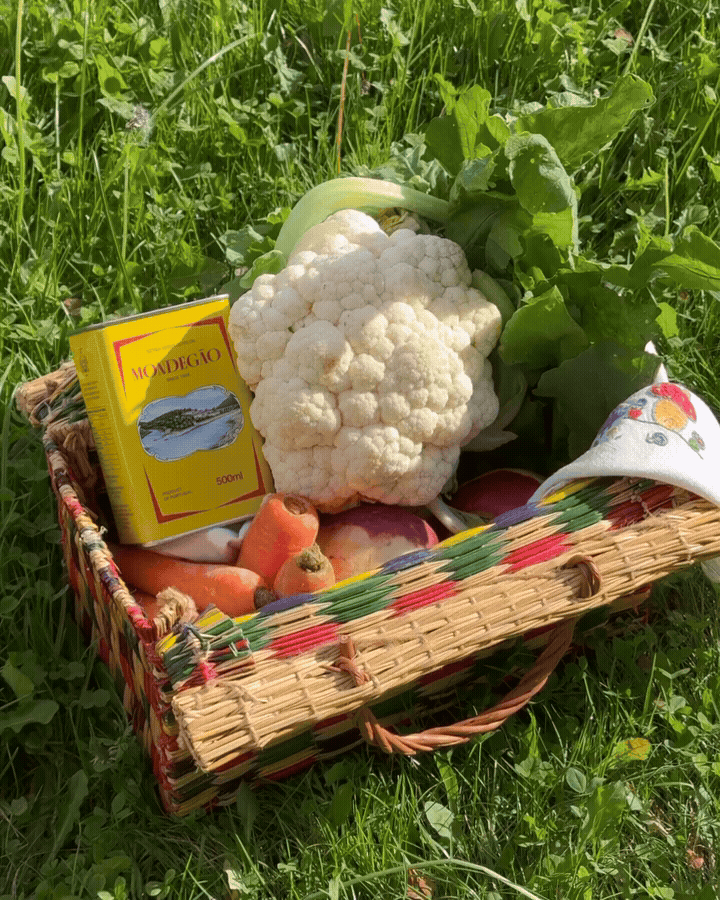 A wicker basket containing a large cauliflower, carrots, eggplants, a box of herbal tea, and a cloth. The basket is on green grass outdoors.