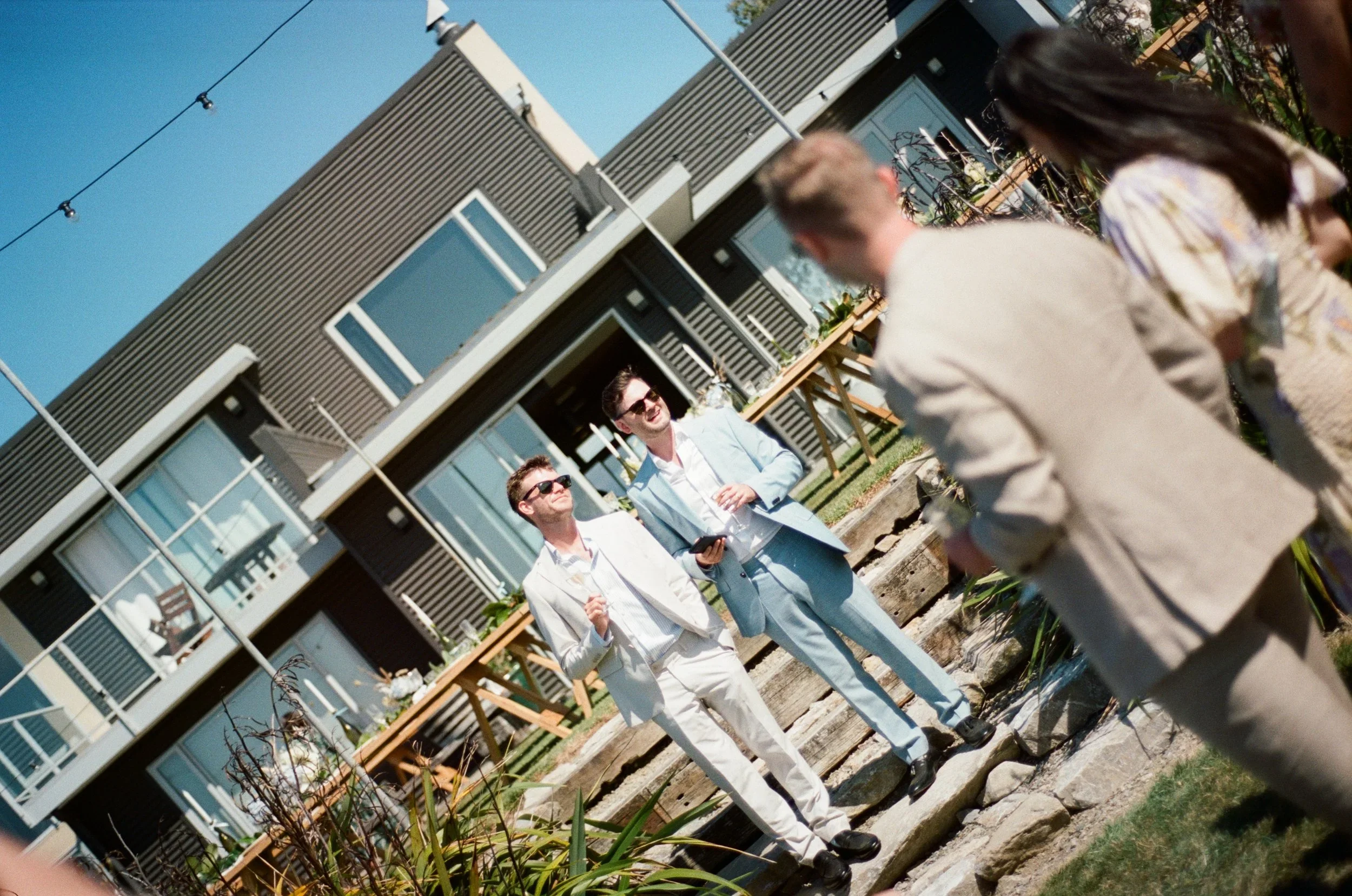 People dressed in suits having a conversation outdoors at a gathering, with a modern building in the background and a table set for an event.