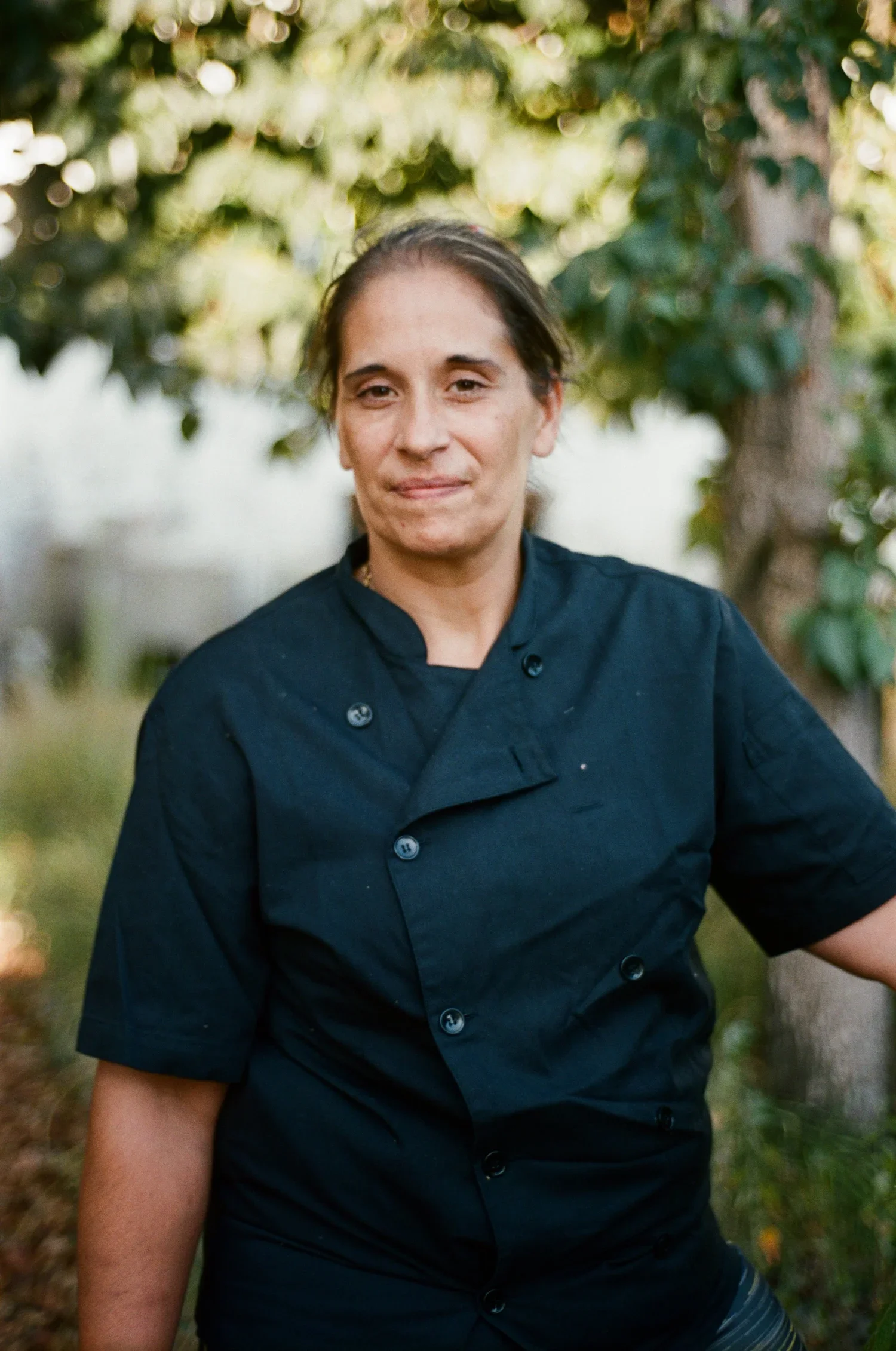 A woman in a black chef's coat standing outdoors with trees and greenery in the background.