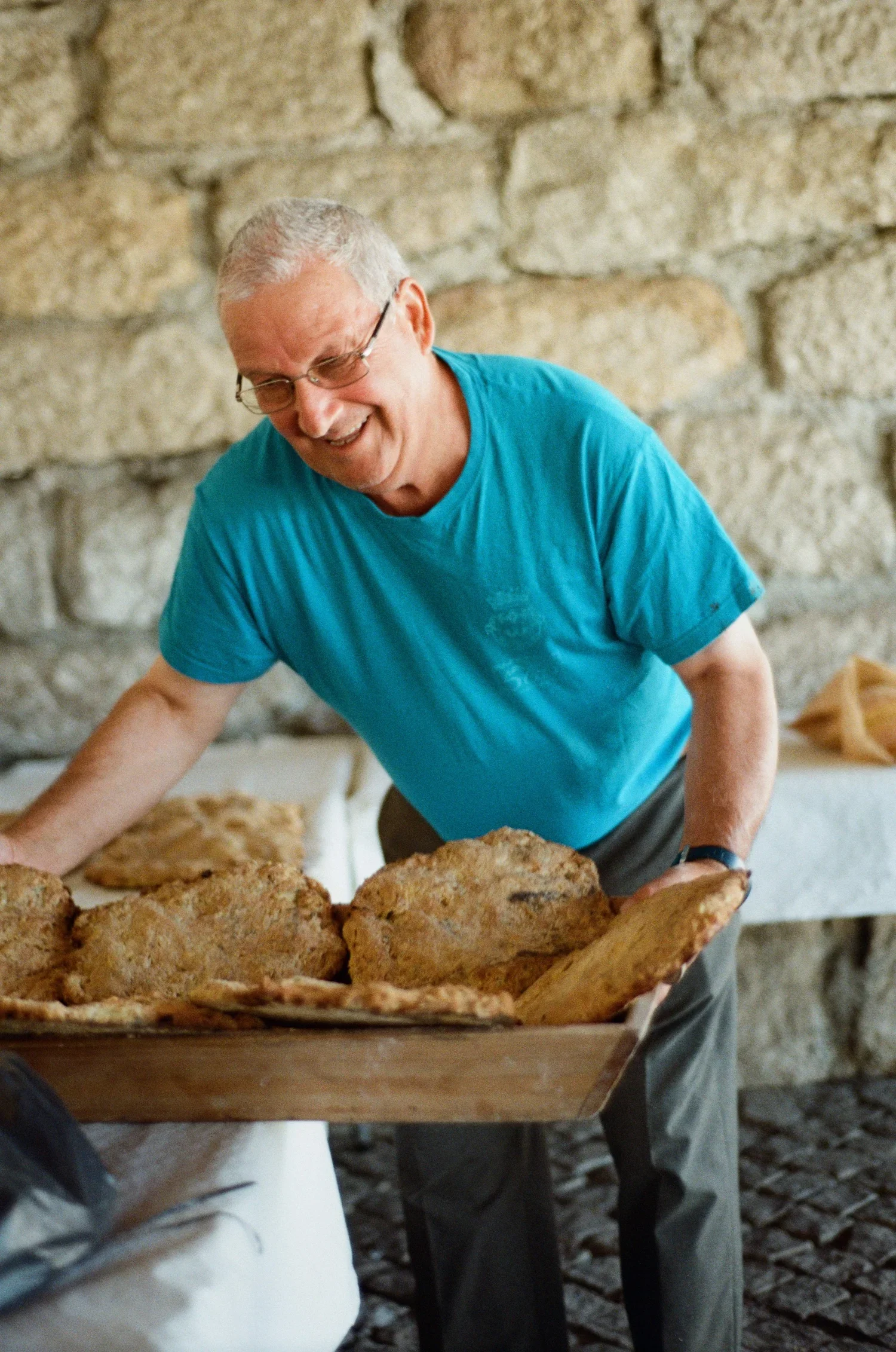 A smiling elderly man with glasses wearing a blue t-shirt, leaning over a tray of freshly baked bread in a rustic stone-walled setting.