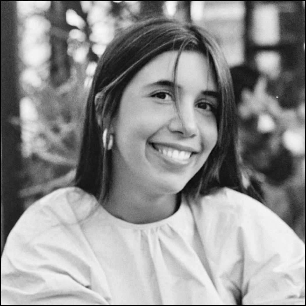 A smiling young woman with long dark hair, wearing a light-colored top and hoop earrings, outdoors with blurred trees in the background.