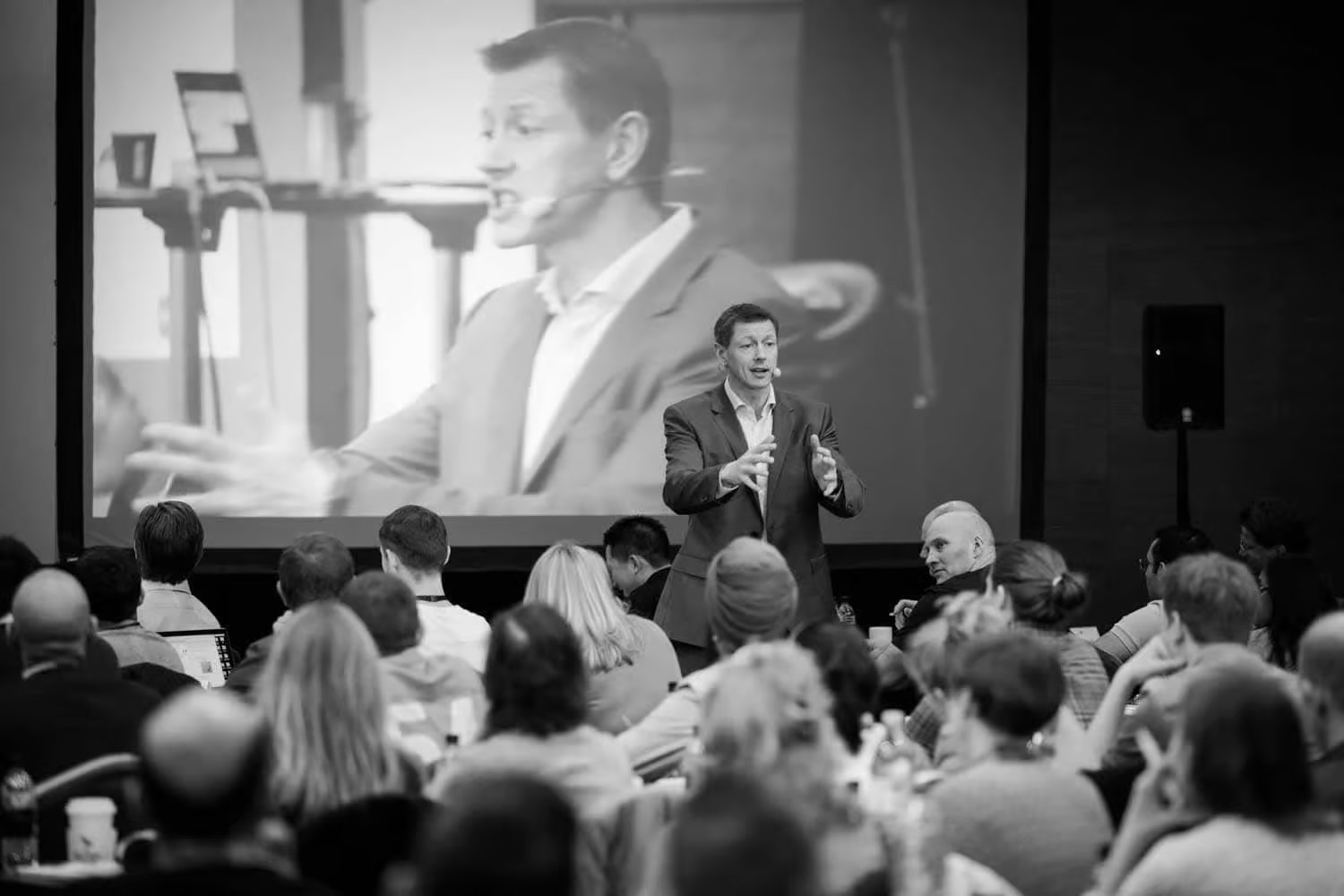 Speaker presenting at a conference with an audience seated and a large screen showing the speaker in the background.