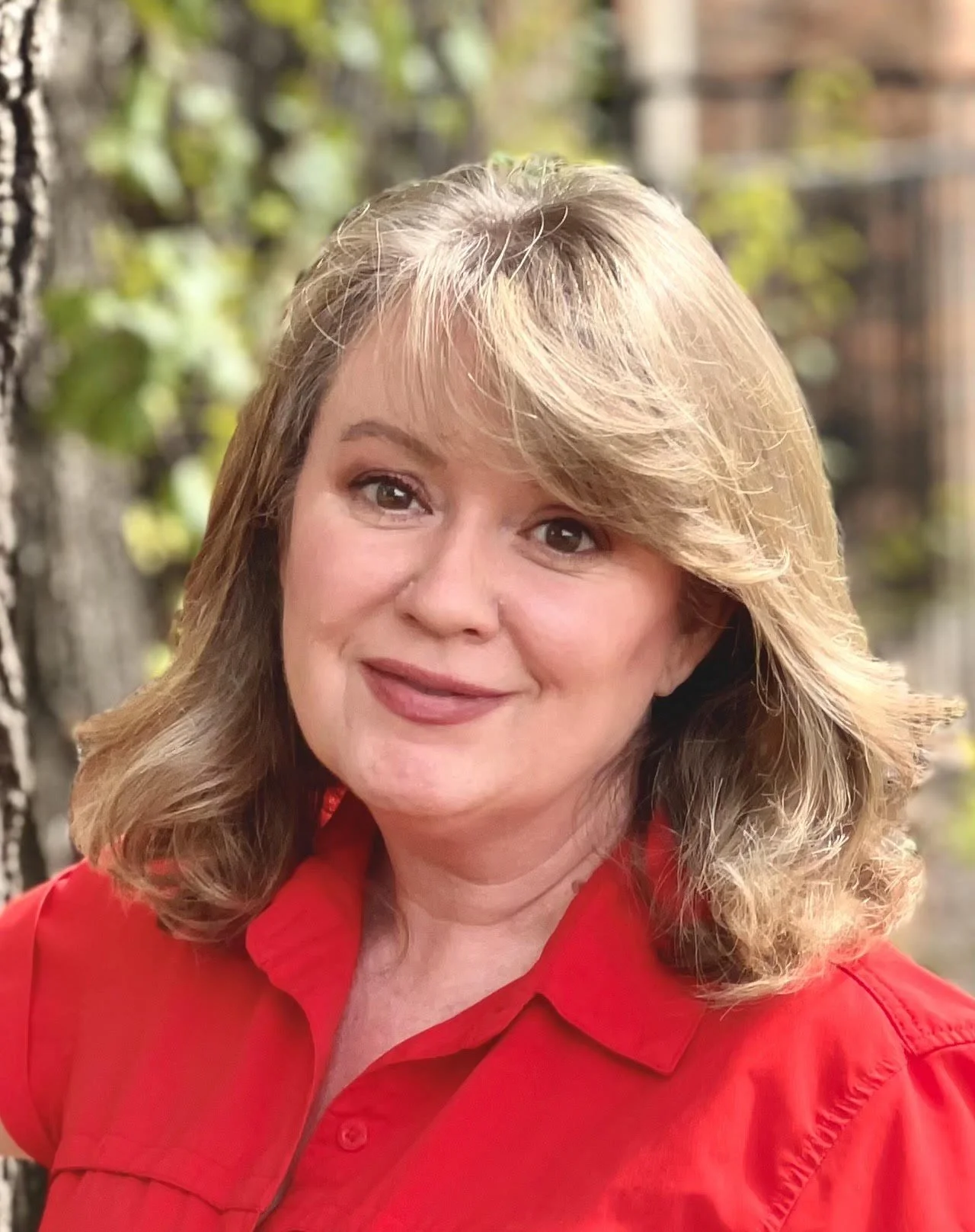 A person with blonde hair wearing a red shirt is smiling at the camera against a blurred outdoor background.