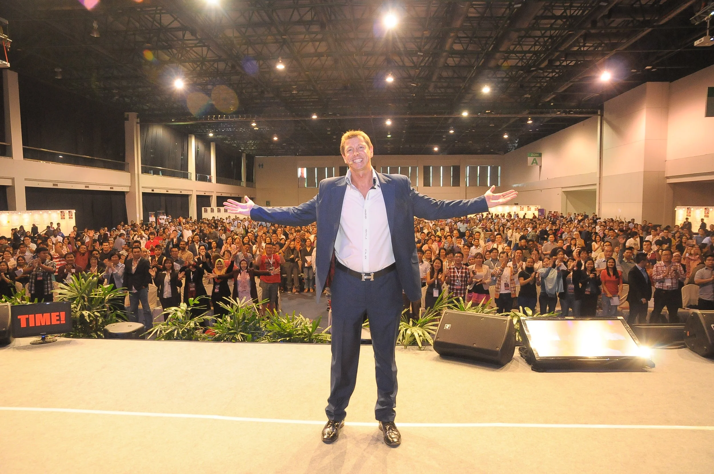 A person in a suit standing on stage with a large audience clapping in a conference hall.