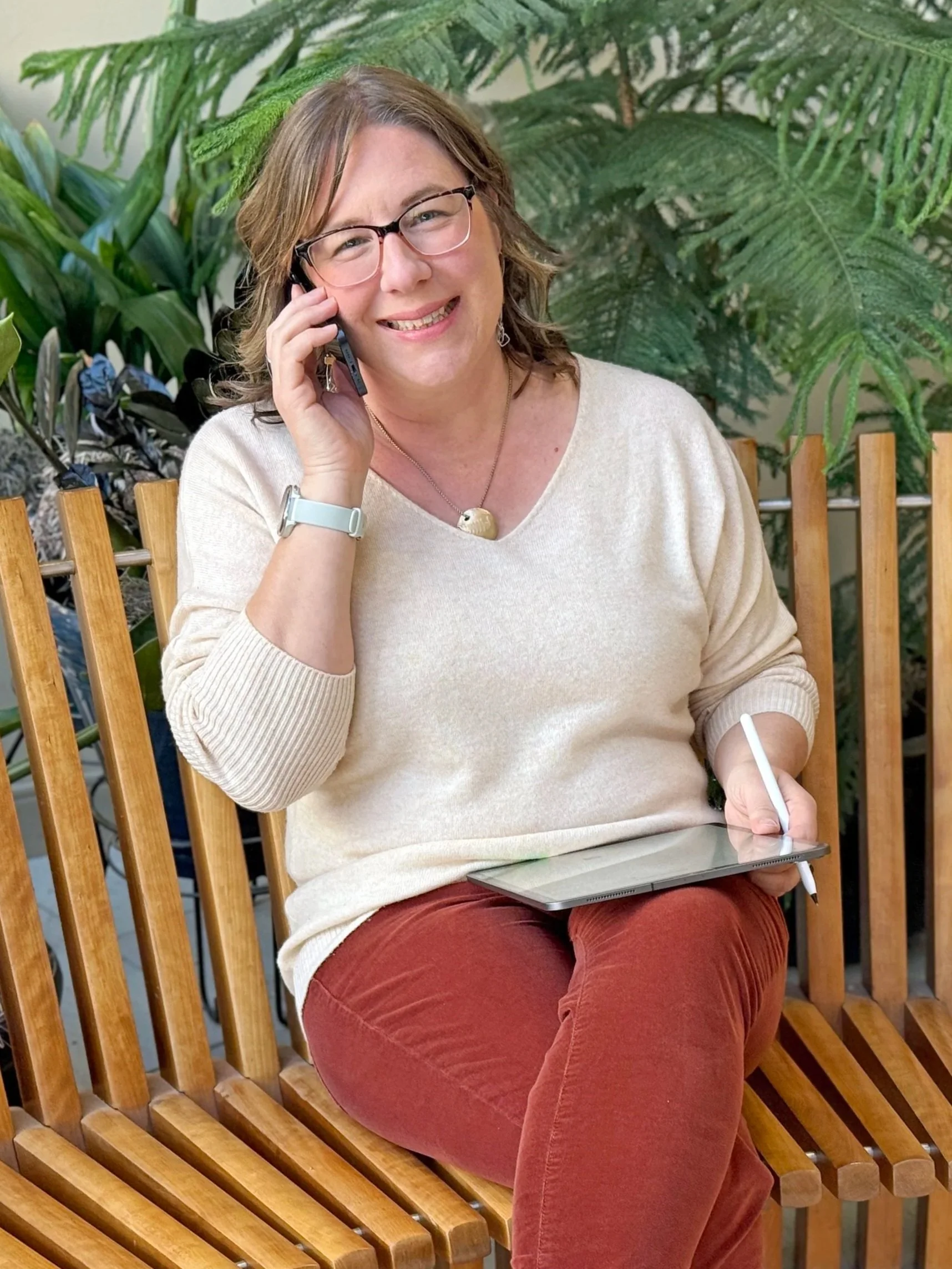 Nonprofit Solution Hub founder Renée Johansson, a woman with shoulder-length brown hair and glasses is sitting on a wooden bench, holding an iPad and Apple Pencil, talking on a cell phone, surrounded by large green leaves and ferns.