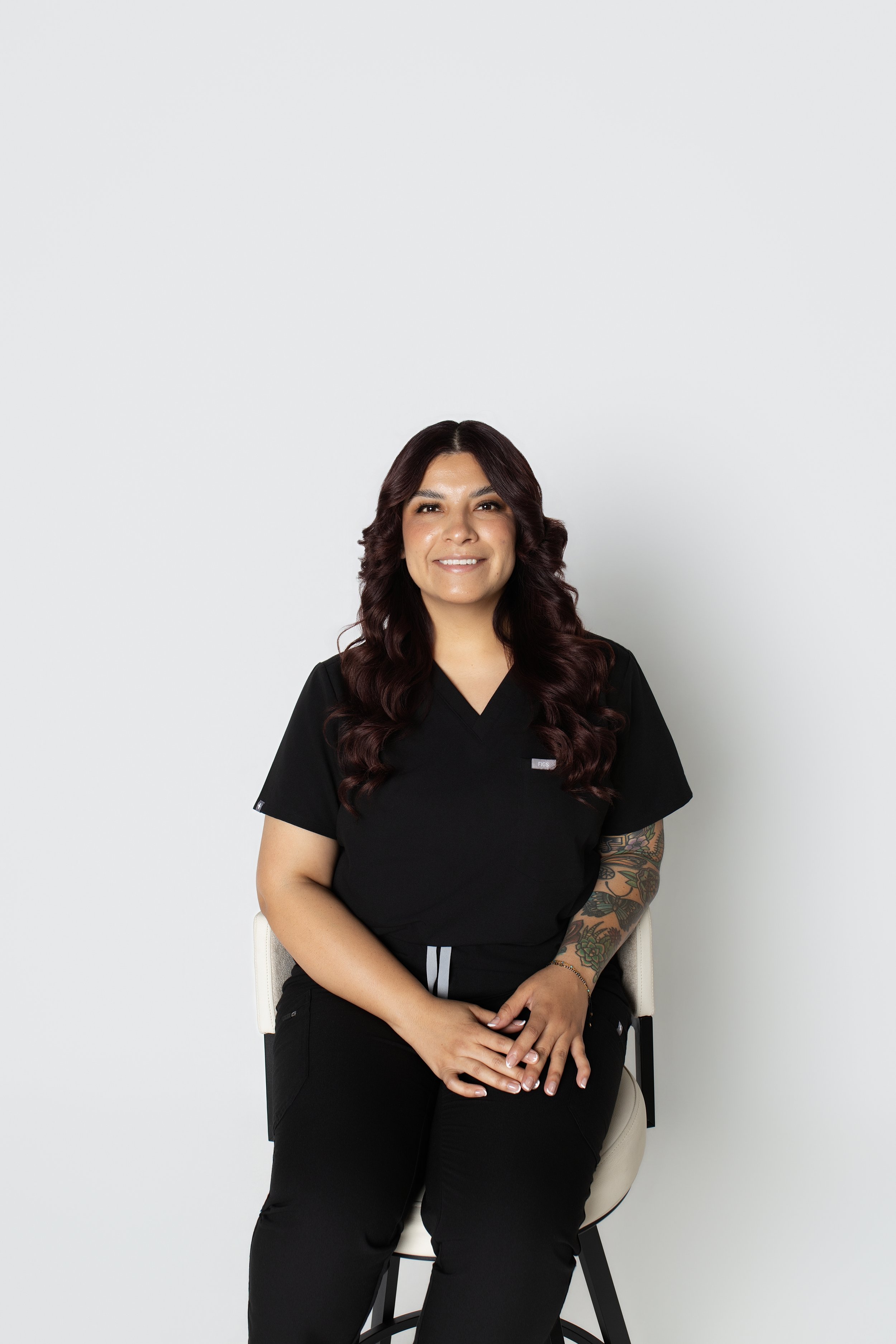 A woman with long, wavy dark hair and tattoos on her left arm, wearing black scrubs, sitting on a white chair with a neutral expression, against a plain white background.