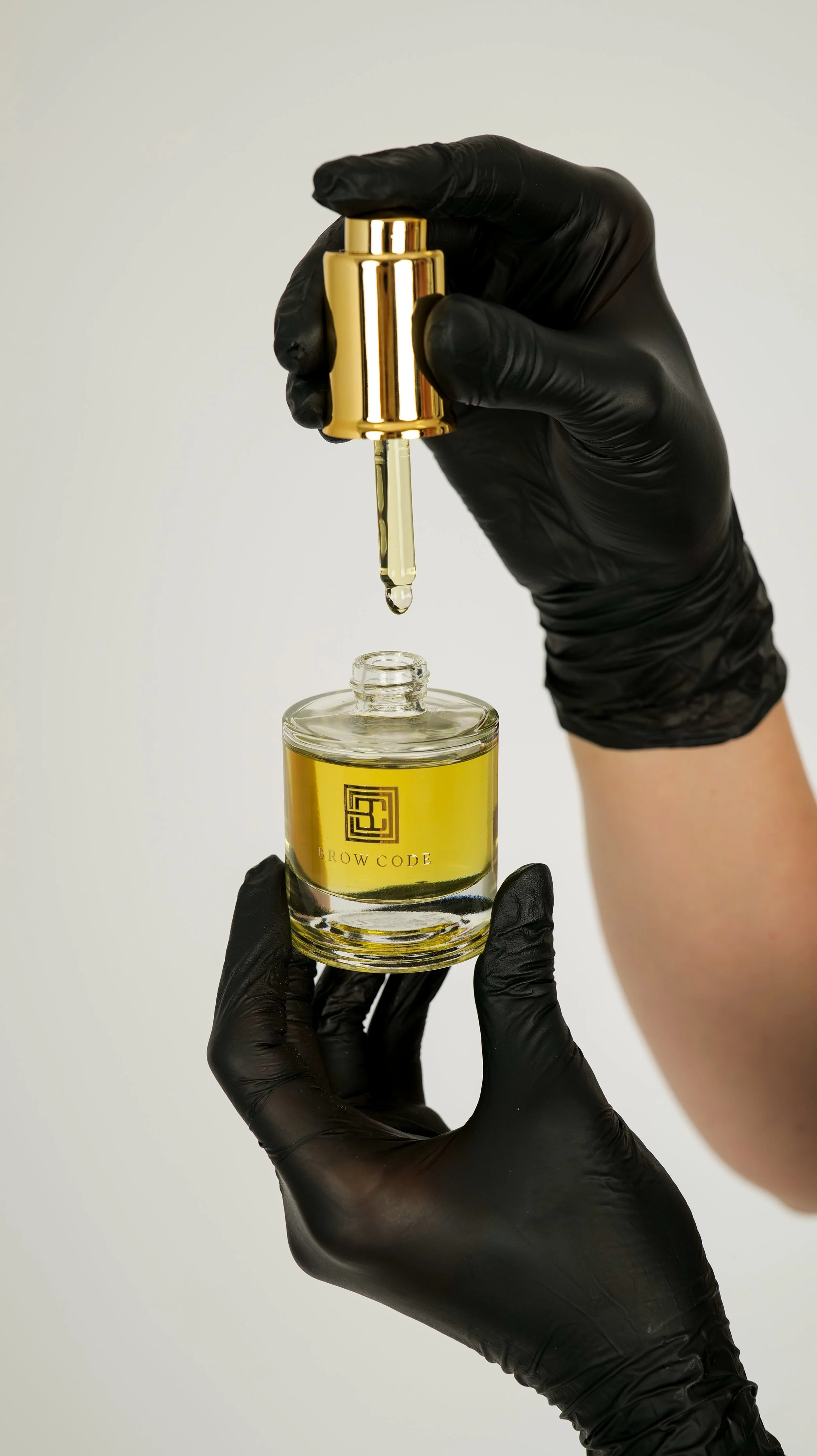 Hands in black gloves holding a gold dropper bottle over a glass jar labeled "BROW CODE" filled with yellow liquid, with a plain light background.