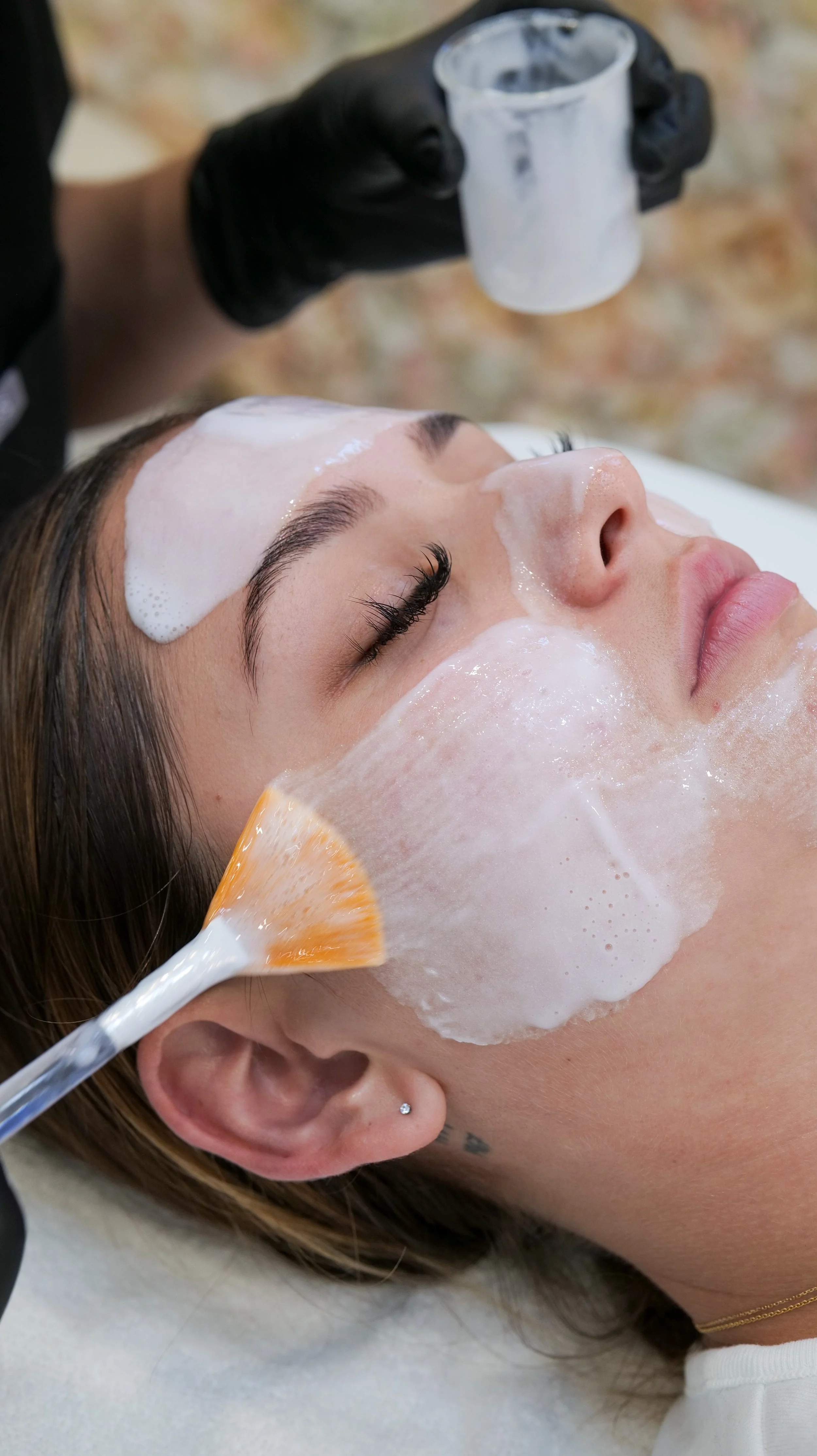 Brightside Esthetics Huntington Beach Estheticians Facials Waxing Chemical Peels Custom Skin Treatments. A woman receiving a facial treatment, with a foam mask on her face. A person with black gloves is applying foam with a brush.