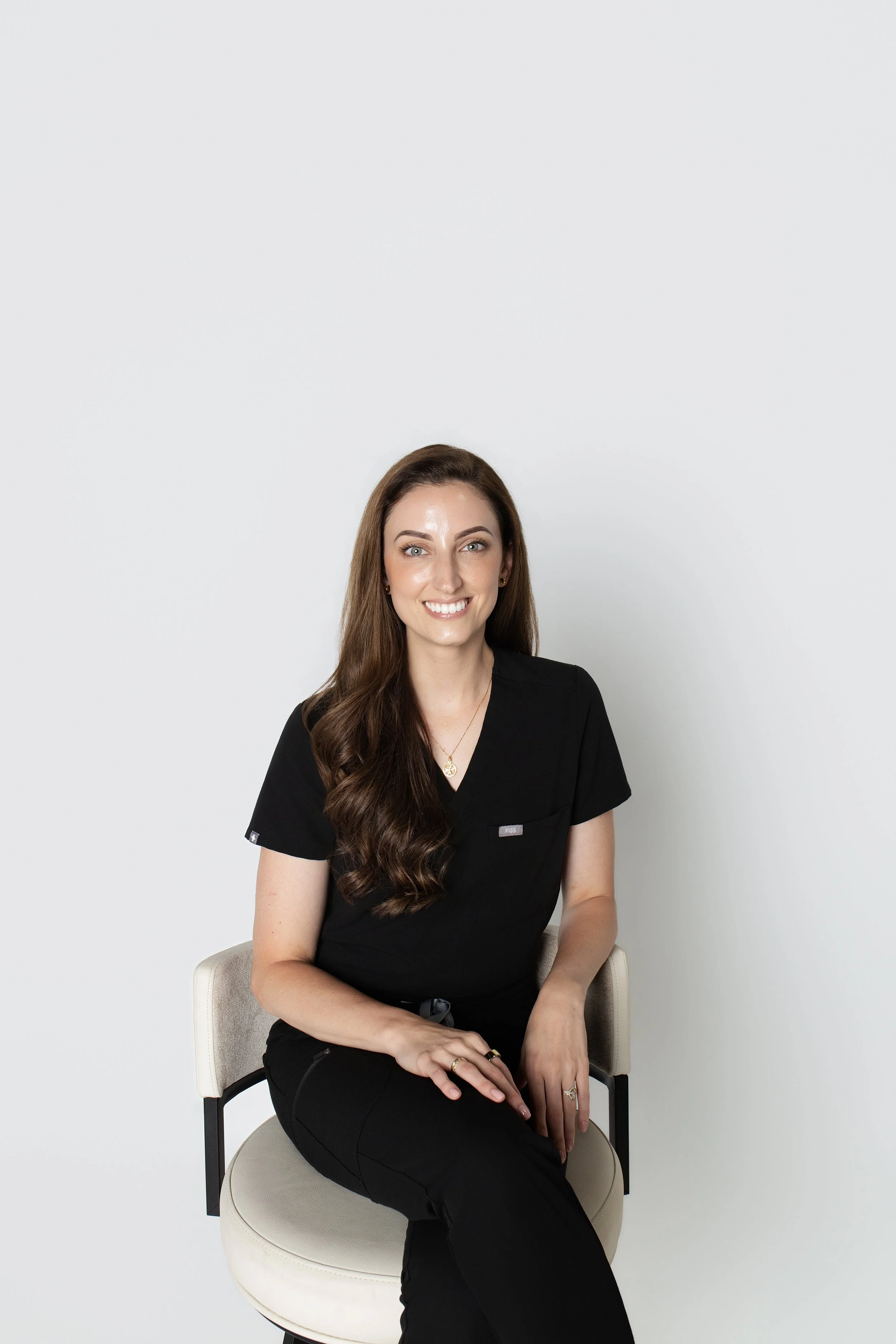 A woman with long wavy brown hair and fair skin sitting on a light beige chair against a plain white background. She is wearing a black top with a V-neck and black pants, adorned with rings and a necklace. She is smiling, looking directly at the camera, with her hands resting on her lap.