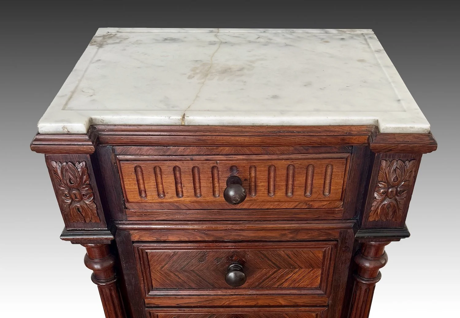 Detail of the white marble top with a stable age crack and molded edge, above carved foliate corner blocks and the fluted frieze drawer.
