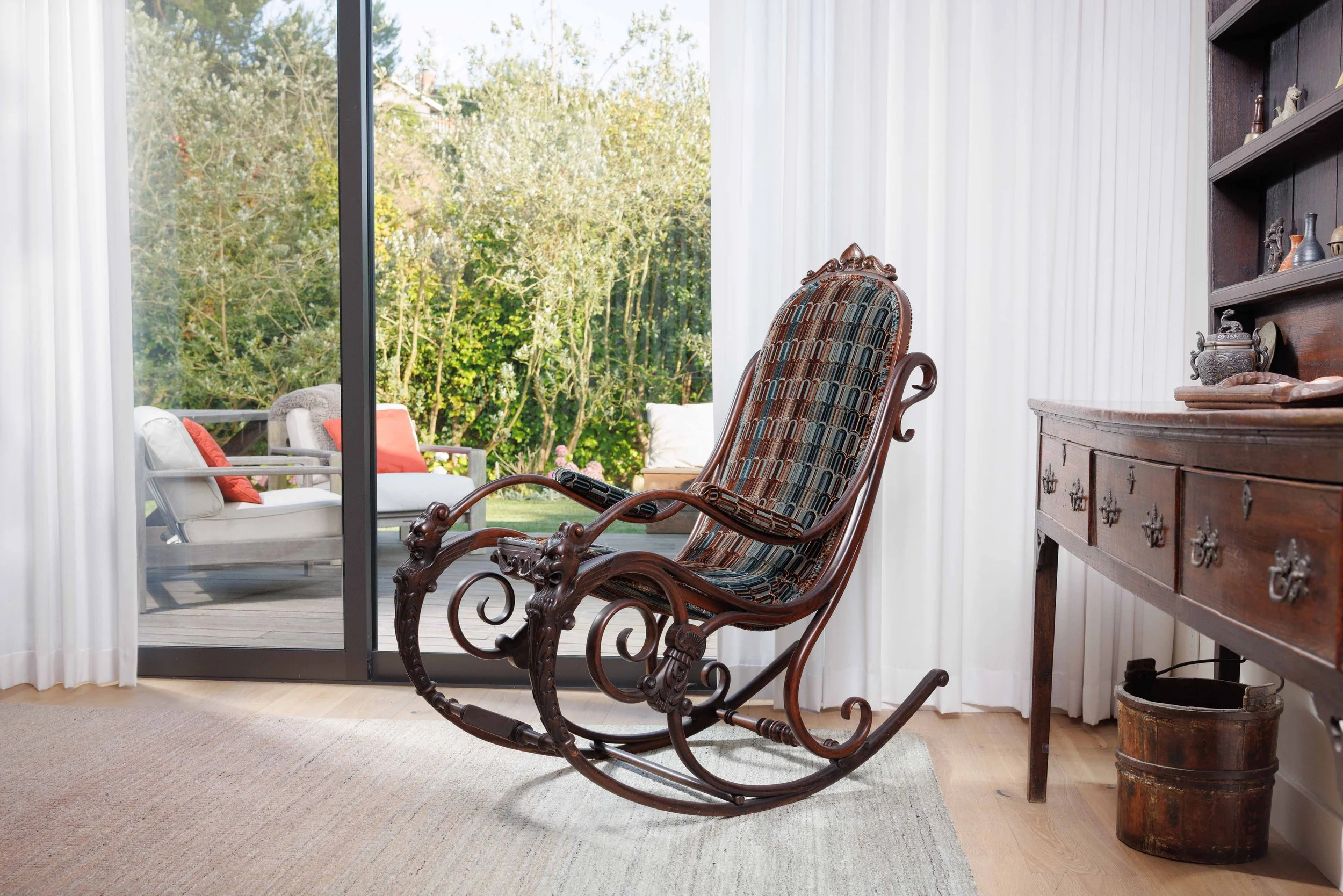 A wooden rocking chair with a colorful patterned cushion inside a room, looking out onto a patio with outdoor furniture and trees outside.