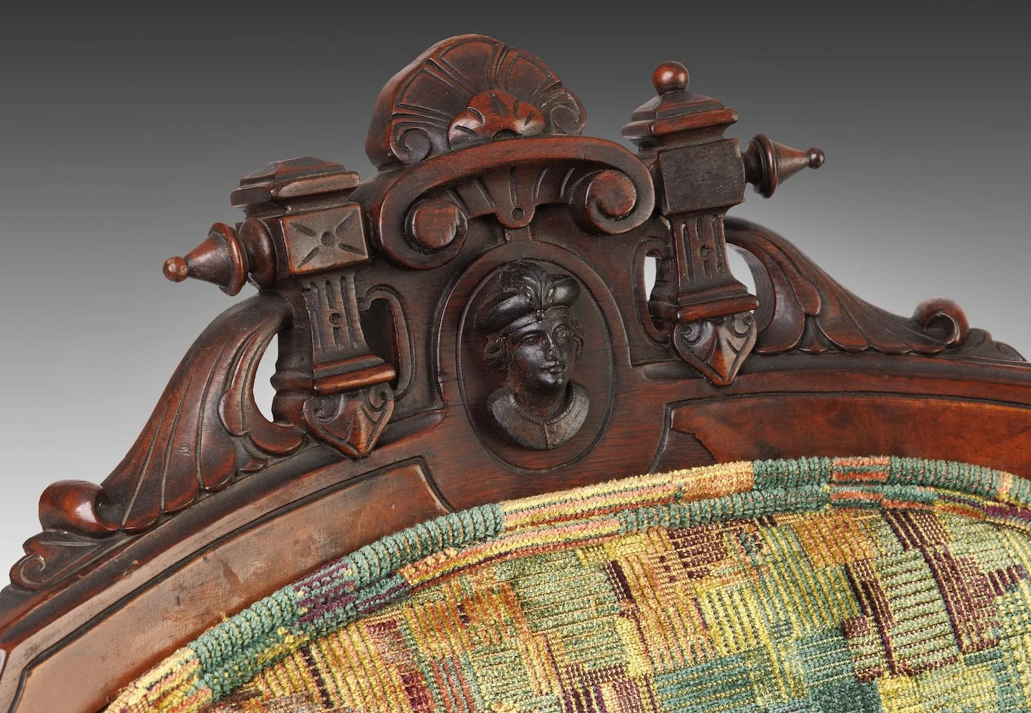 Carved walnut crest of a parlor chair, its oval medallion holding a man's bust in a plumed cap, flanked by turned finials and scrollwork, above checkerboard velvet