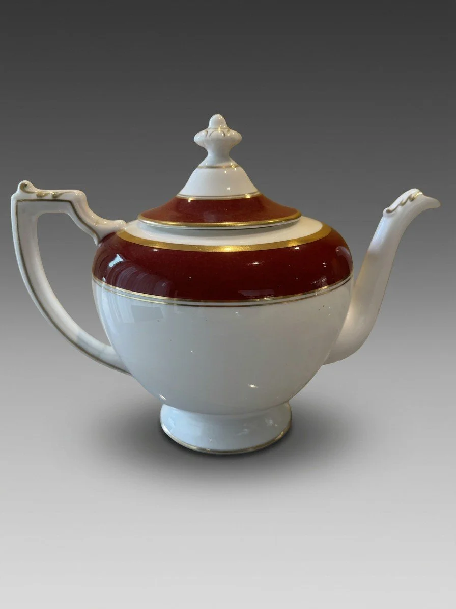 A red and gold bone china teapot photographed from the side, lid in place, the handle turned toward the camera.