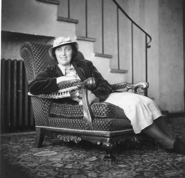 Woman seated in a carved scroll-arm chair upholstered in patterned velvet, a staircase rising behind her, interior portrait circa 1935