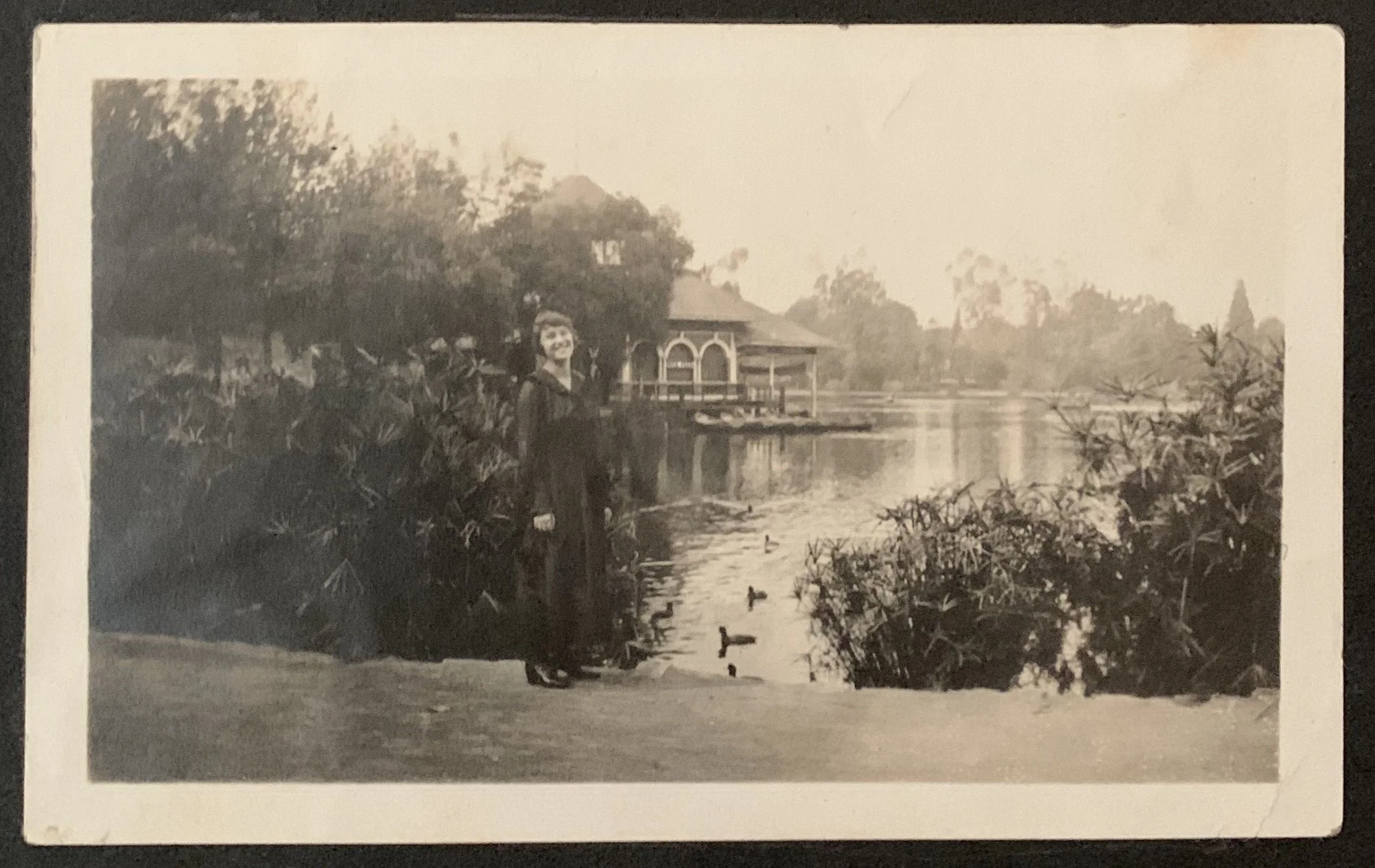 Early twentieth century photograph of a young woman standing at the edge of the lake in Westlake Park, the boathouse and ducks on the water behind her