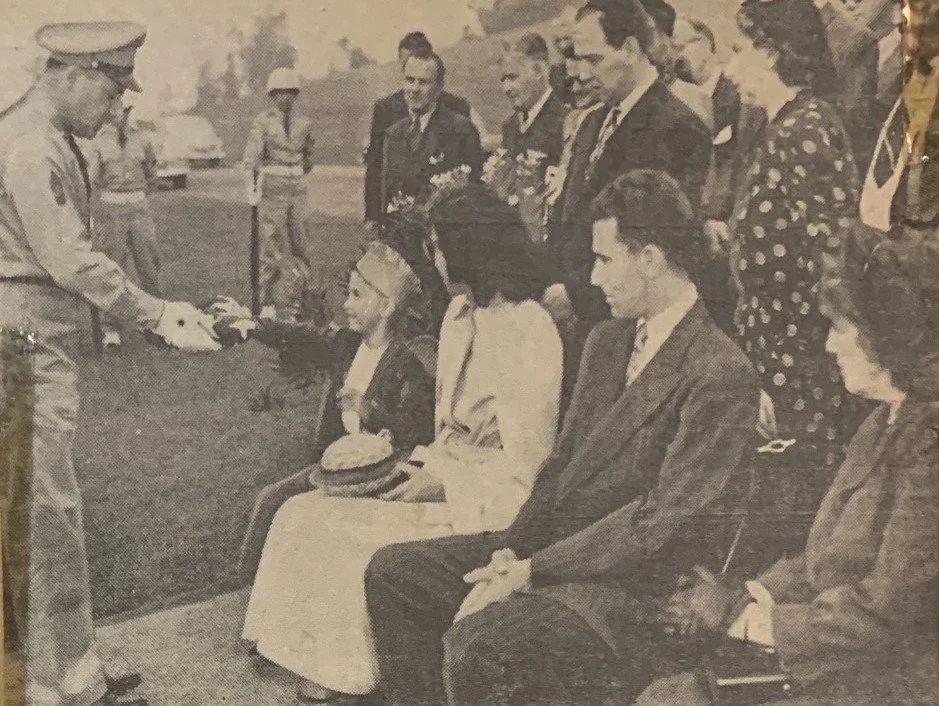 A uniformed officer in white gloves presents a folded American flag to a young boy seated with family at a military funeral, about 1949.