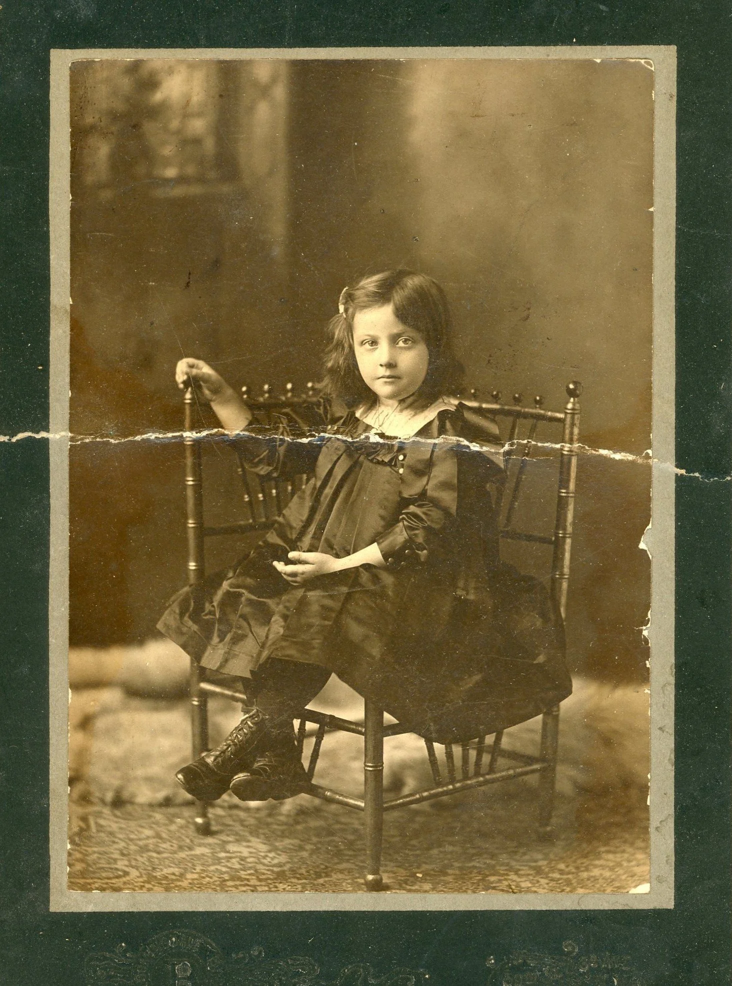 Young girl in laced boots seated in a turned-wood spindle-back chair, studio portrait circa 1902, a patterned rug and pale fur at her feet