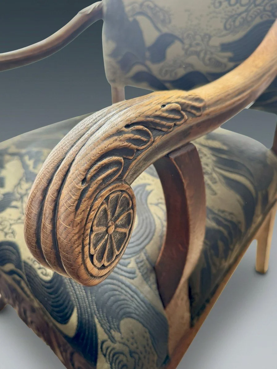Detail of the Solstice Crest carved oak arm, showing a leaf-and-rosette volute and the deepened tone of wear along the outer curve