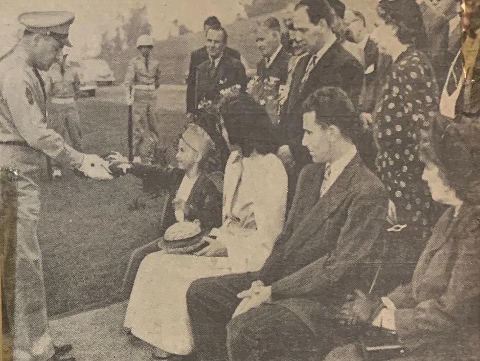 A uniformed officer in white gloves presents a folded American flag to a young boy seated with family at a military funeral, about 1949.