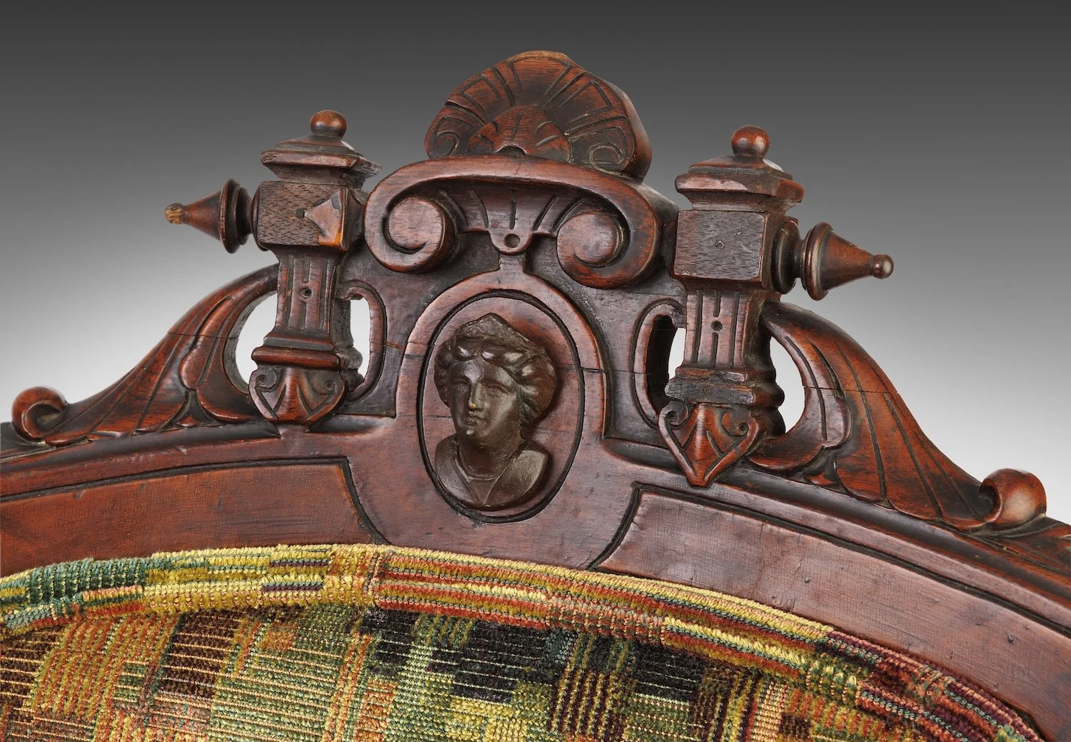 Carved walnut crest of the companion chair, its oval medallion holding a woman's bust with bound hair and a wreath, above checkerboard velvet