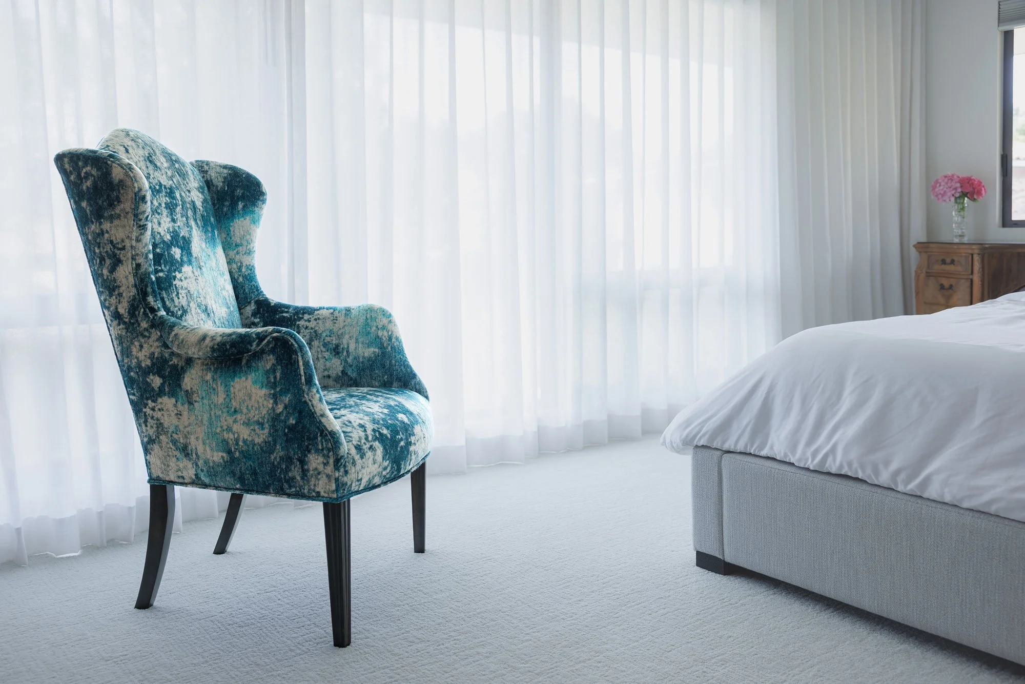 The restored wingback chair in mottled teal and ivory velvet stands on pale carpet against sheer curtains, beside the foot of a bed.