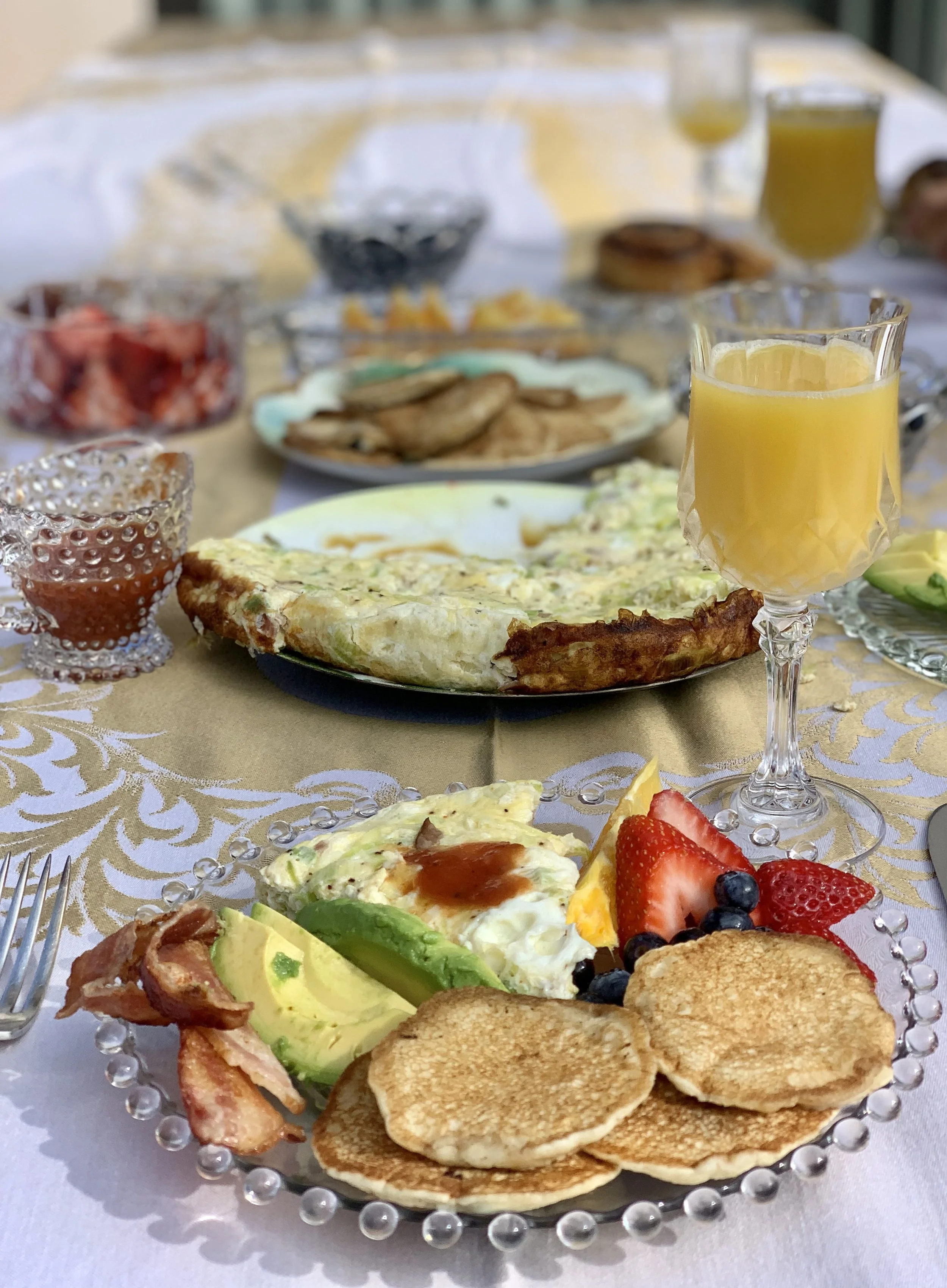 A breakfast of pancakes, bacon, egg, and fruit on a Candlewick beaded-rim glass plate, with a cut-crystal footed goblet of juice on a damask cloth