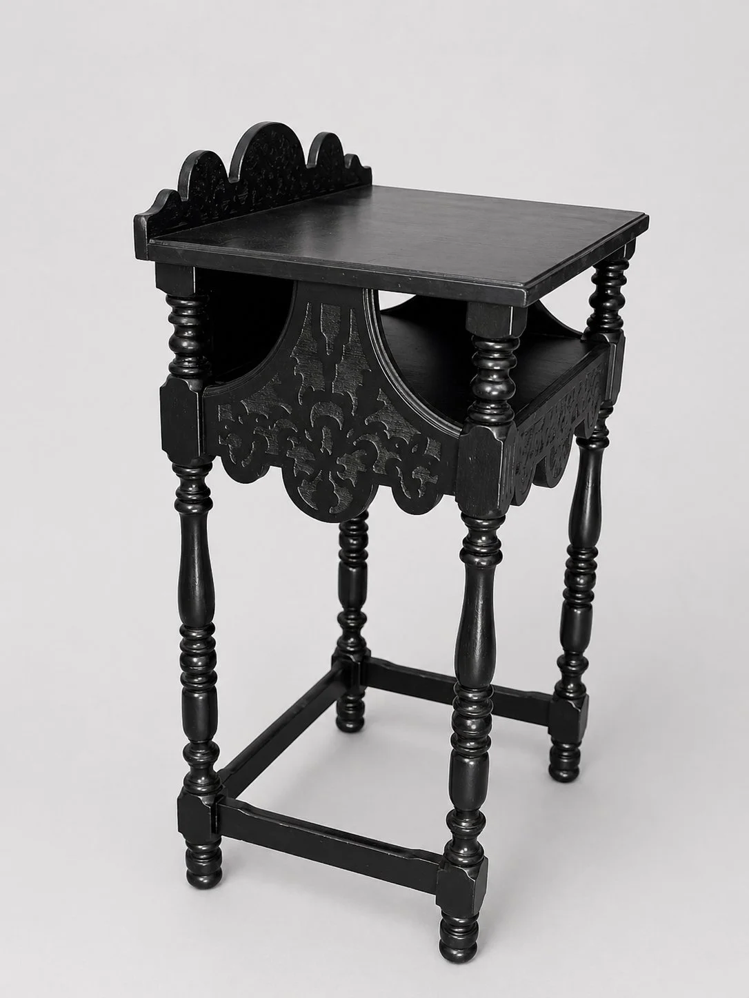 Refinished 1920s telephone stand in black with turned legs, scalloped crest back, and pierced apron, shown in angled front view