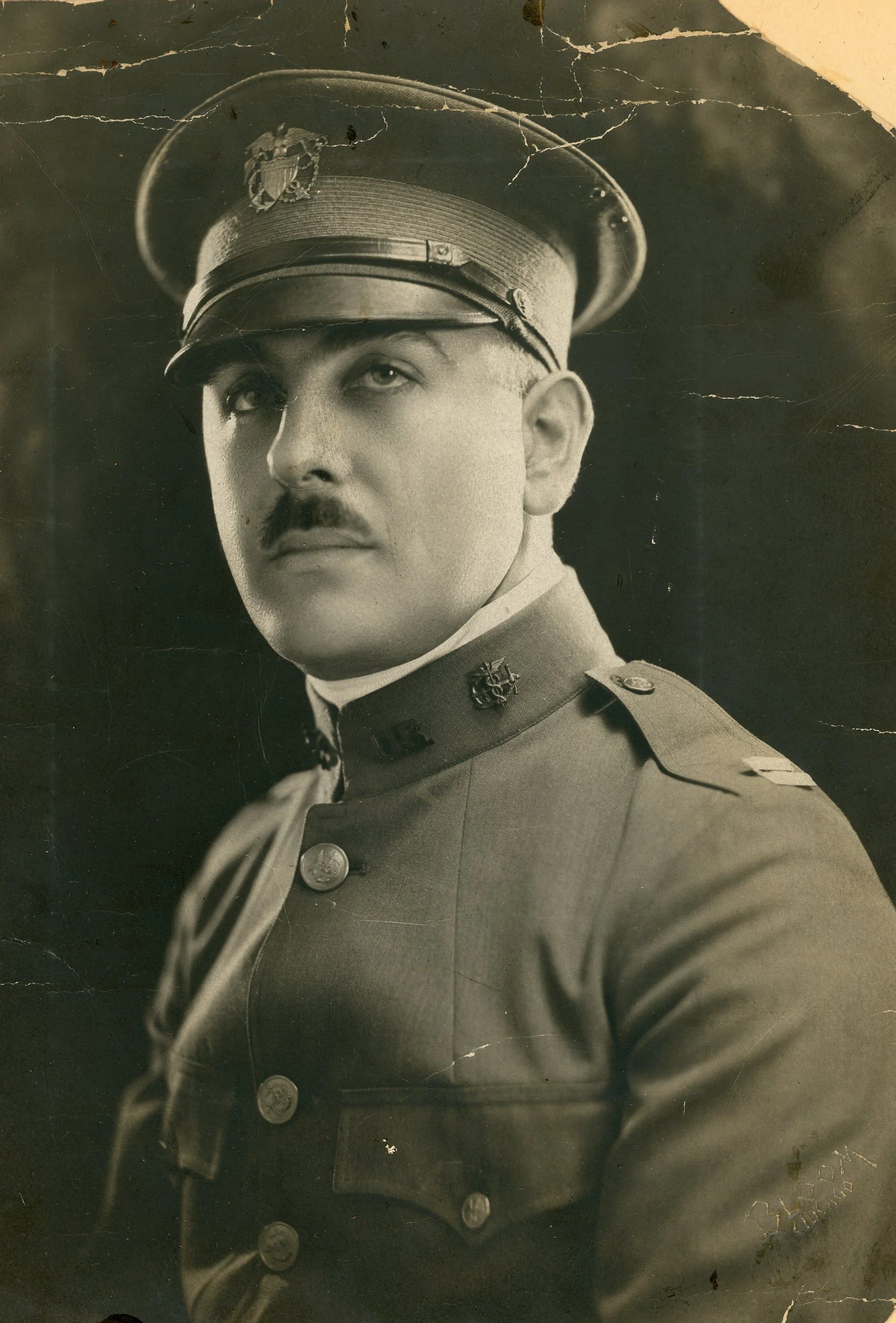 Sepia studio portrait of a young man in a buttoned olive-drab officer's service coat, facing the camera with his shoulders square.