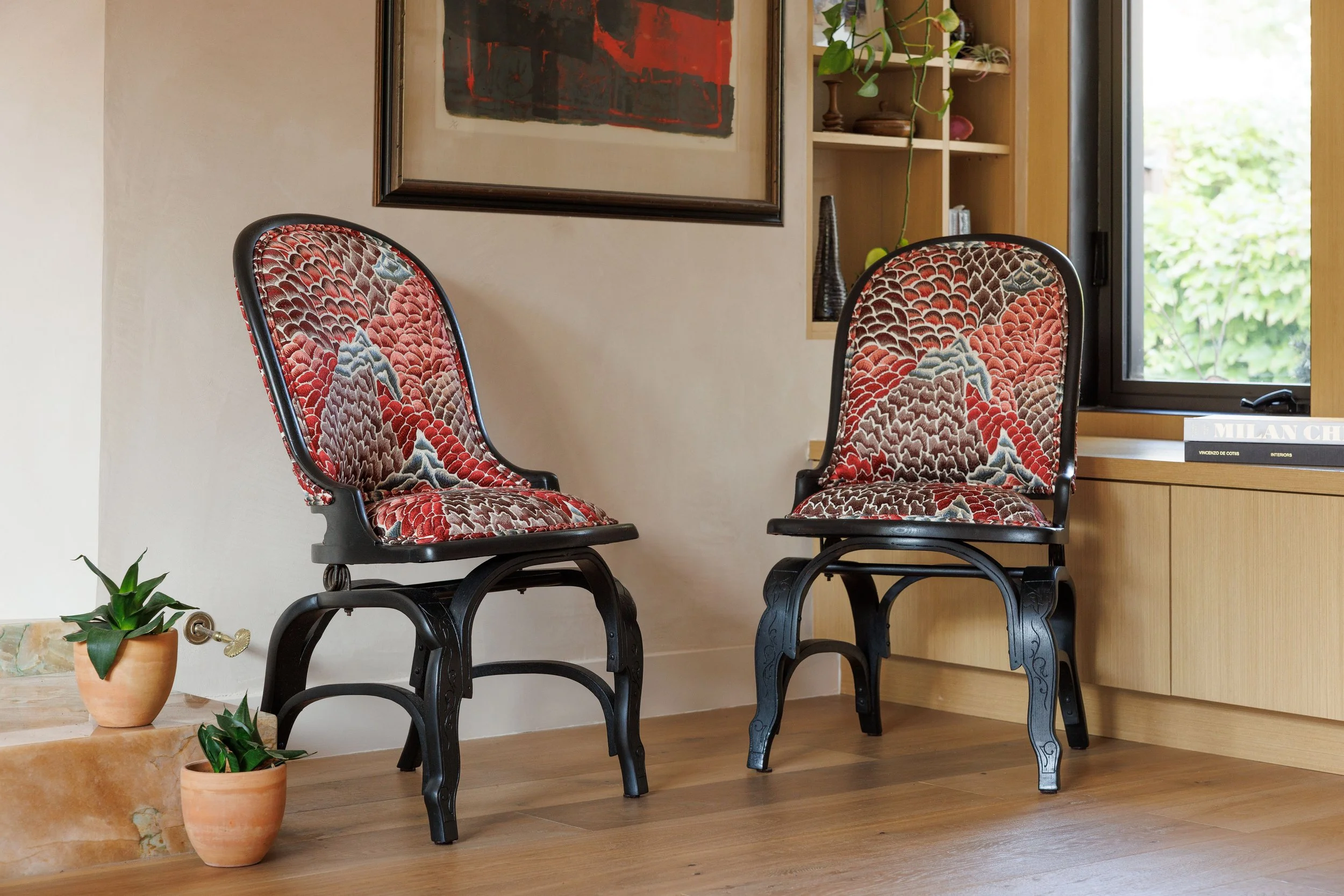 Two vintage-style chairs with upholstered cushions featuring a colorful bird and leaf pattern, placed in a room with wooden flooring, a window with a view of green foliage, and decorative plants around.