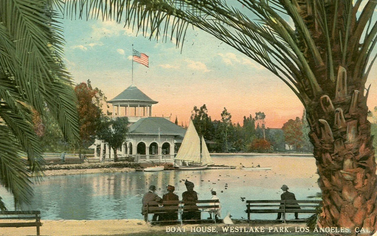 Hand-tinted postcard of the boathouse at Westlake Park Los Angeles, with figures seated on wooden benches at the water's edge beneath a palm