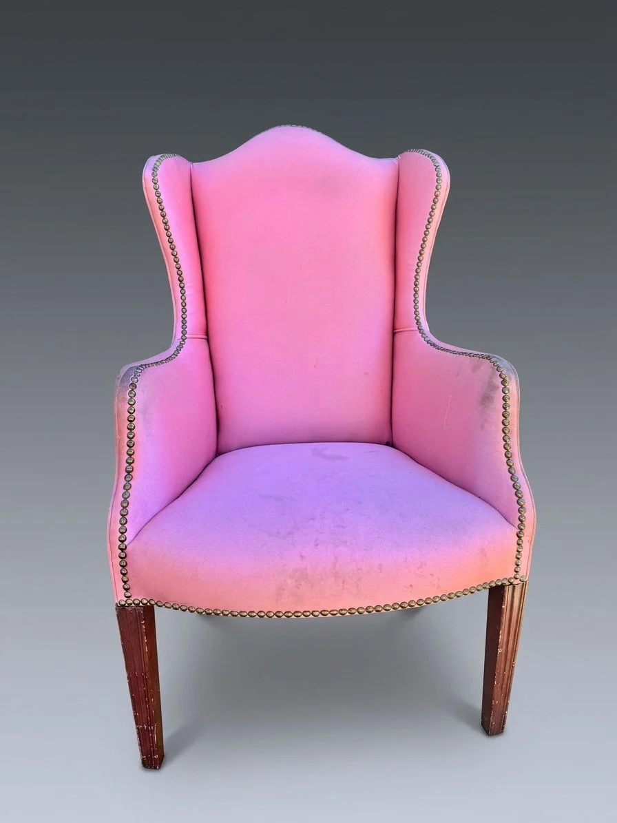 The same wingback chair before restoration, in a soiled pink covering with brass nailhead trim, its fluted front legs worn and dull.