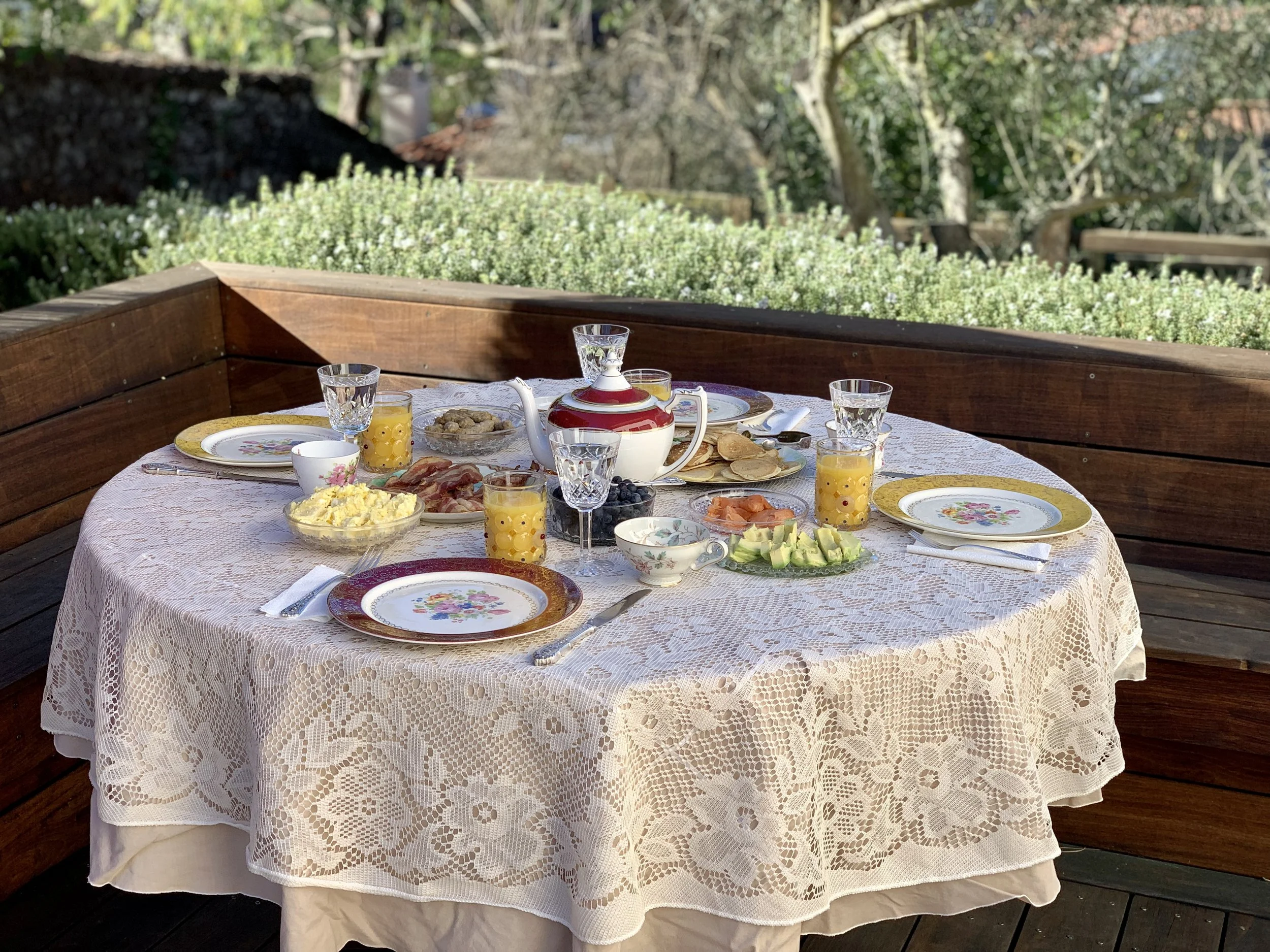 Round outdoor table on a wooden deck set with mismatched gilt-rimmed china, cut glass, and a red-and-gold teapot on a lace cloth.